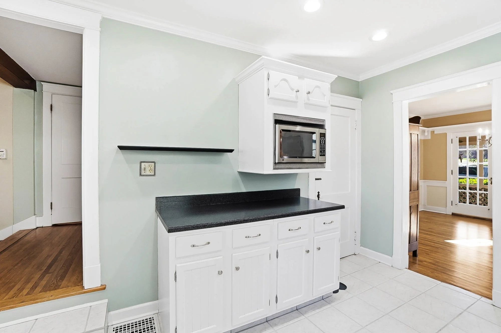 Kitchen corner with white cabinets, black countertop, microwave, and doorway to an adjacent room with wood floors.