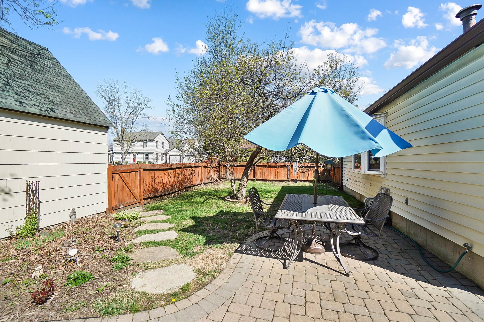 Backyard patio with a round table, four chairs, and a large blue patio umbrella. There is a tree with budding leaves, a grassy area, a stone pathway, and a wooden fence. The house has yellow siding, and neighboring houses are visible in the backgroun