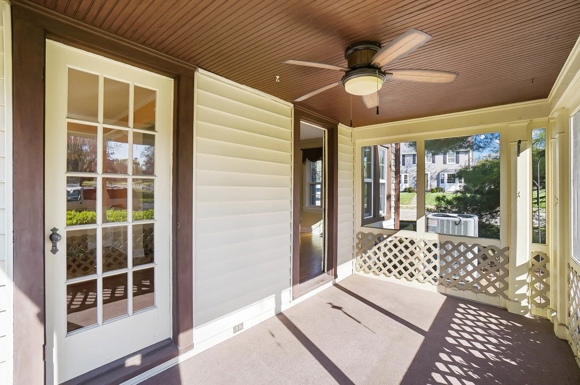 A porch with a ceiling fan, lattice railing, and a partially open screen door leading inside a house. There are large windows, and the porch is bathed in sunlight.