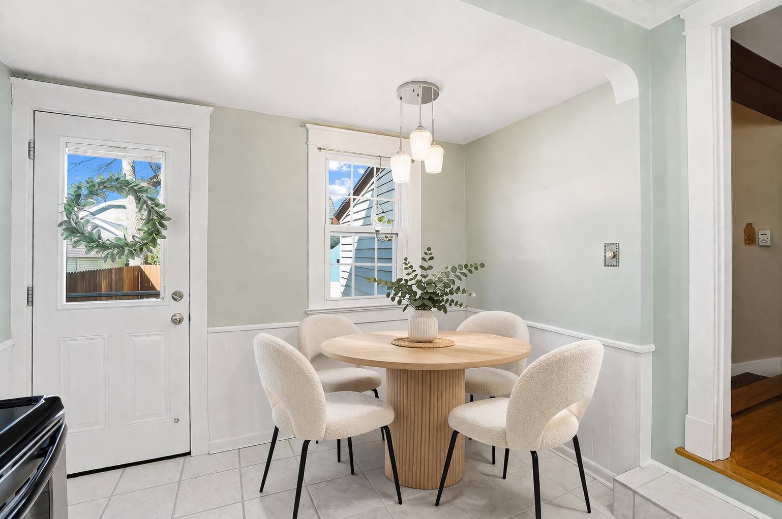 Dining area with a round wooden table, five beige upholstered chairs, a white vase with green eucalyptus leaves, a window with blue sky and house outside, and white walls with wainscoting.