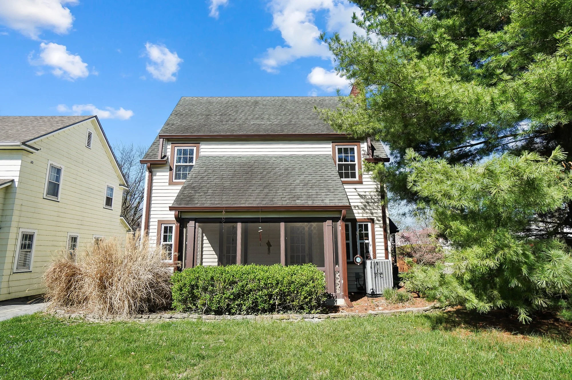 A two-story house with a screened porch, surrounded by green yard and trees, under a blue sky with some clouds.