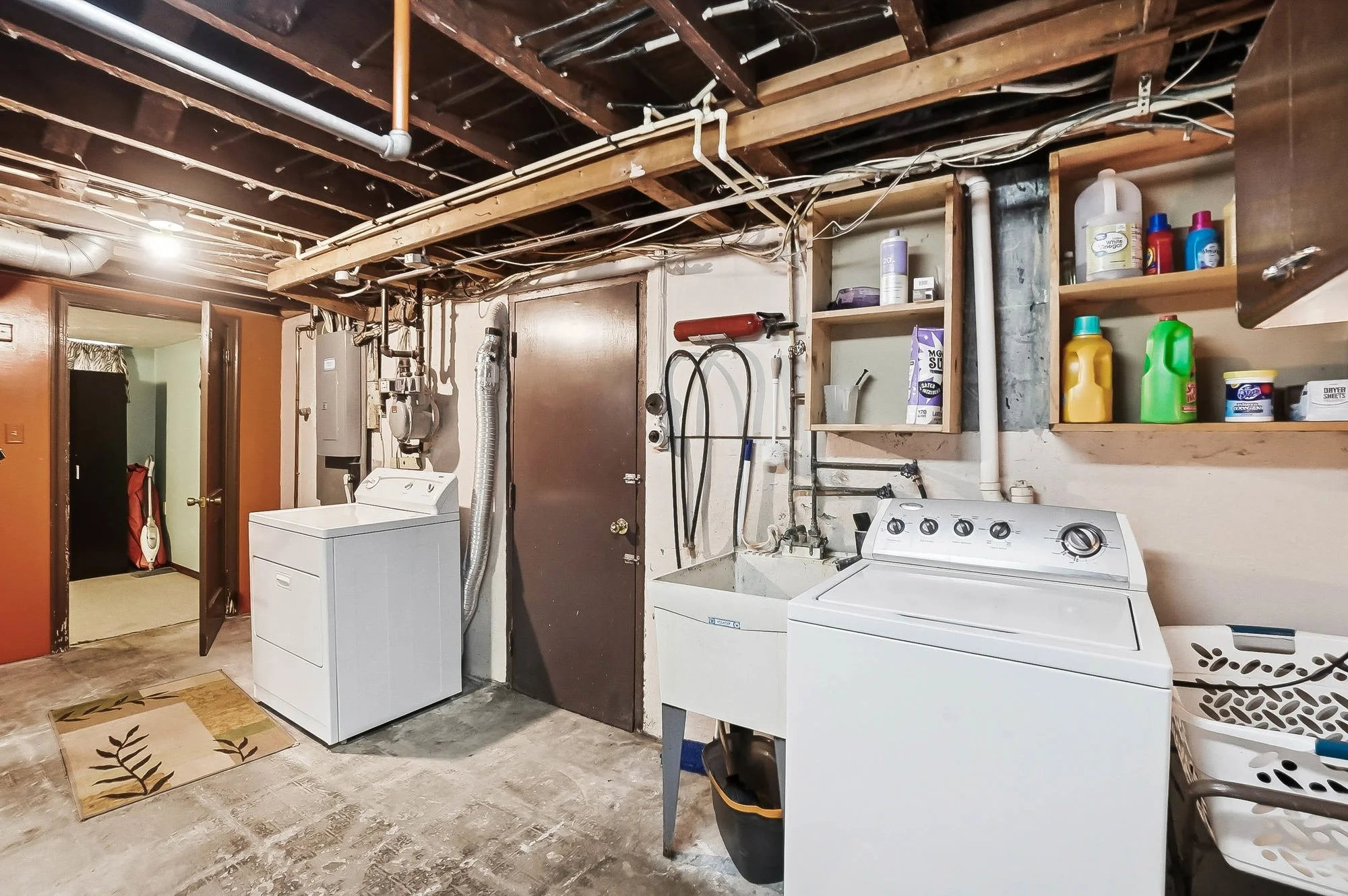 A laundry room with a washing machine, a utility sink, shelves with laundry and cleaning supplies, and exposed ceiling pipes and beams.