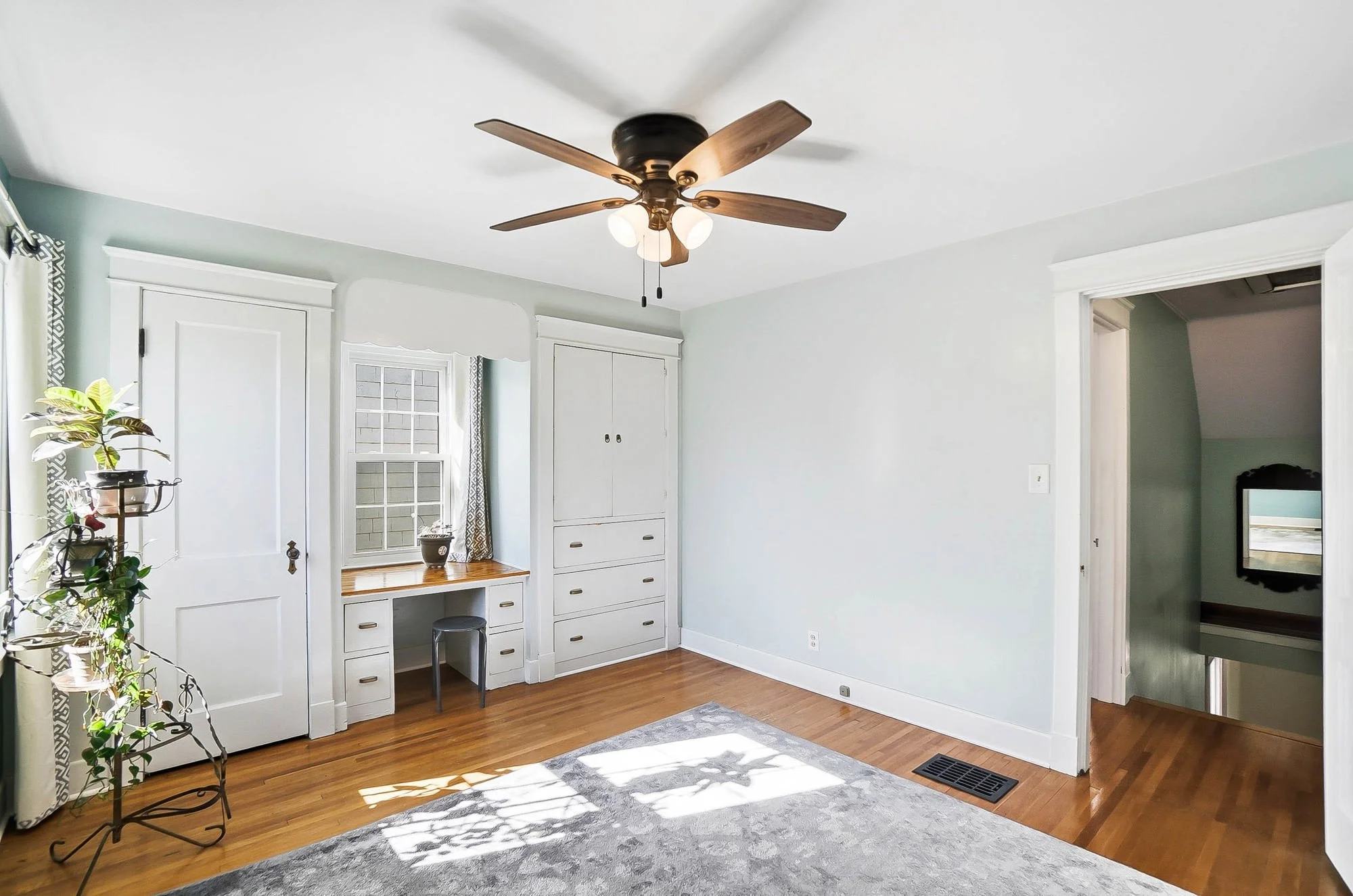 Empty room with hardwood floors, white walls, ceiling fan, built-in storage, window with curtains, and a doorway leading to another area