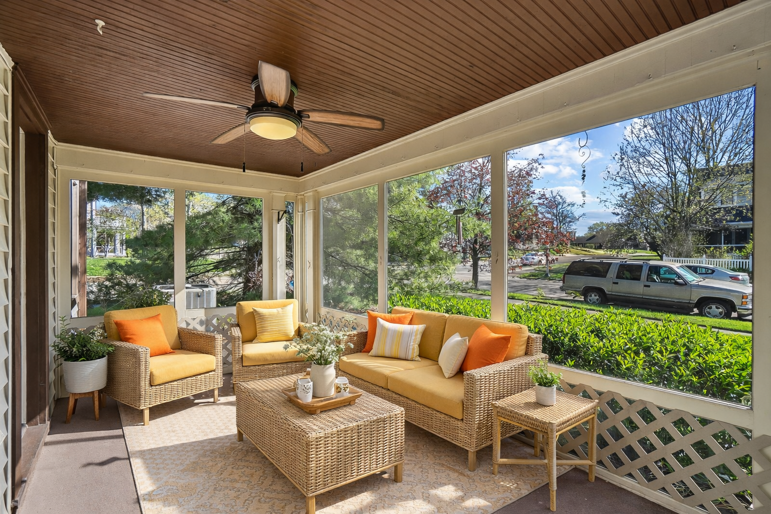 Sunroom with wicker furniture, yellow cushions, and potted plants, overlooking a street with trees and parked cars.
