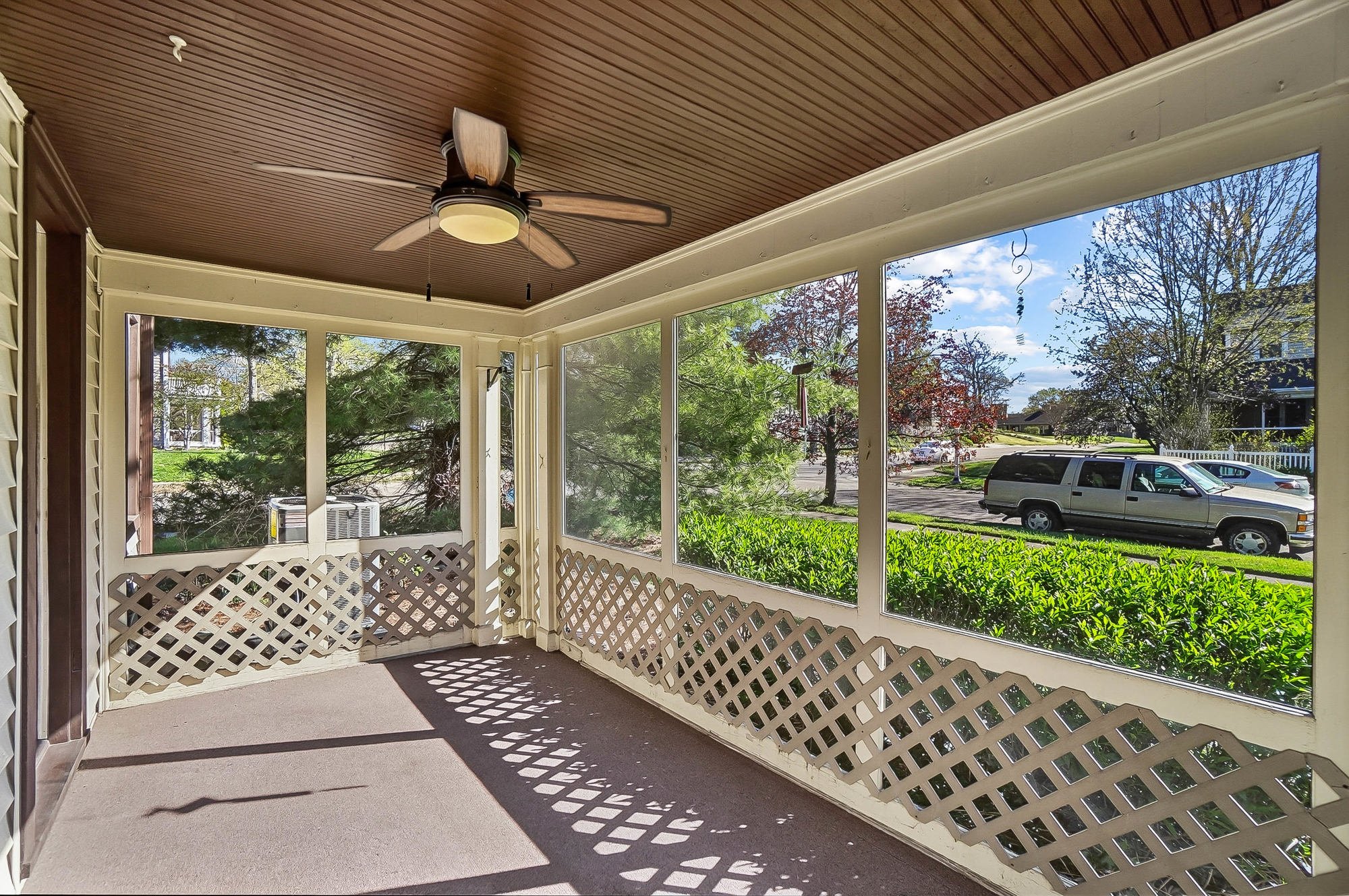 A screened porch with a wooden ceiling, ceiling fan, and lattice panels, overlooking a lawn, trees, and a street with parked cars.