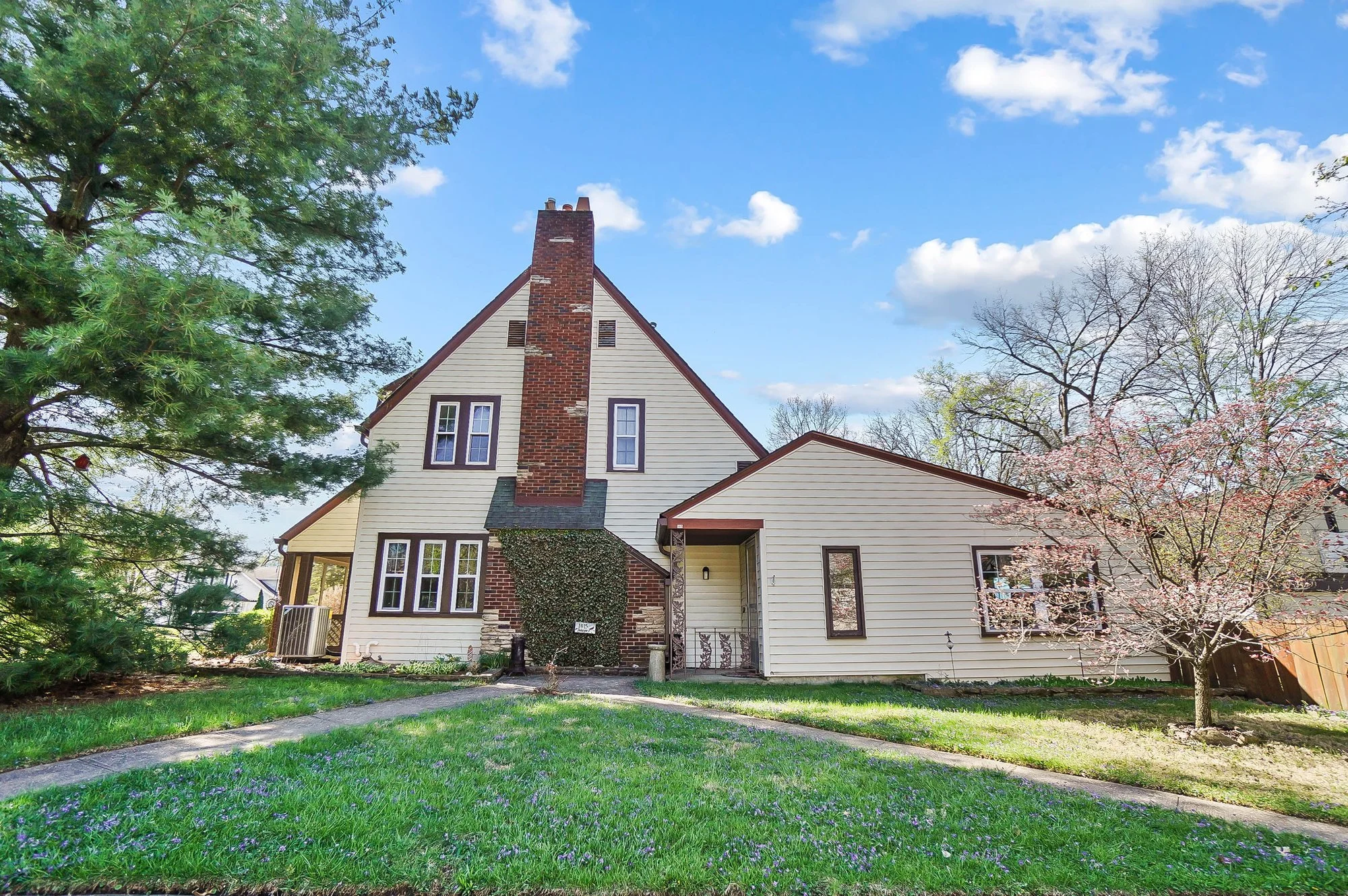 A two-story house with a brick chimney, beige siding, and a side porch, surrounded by green grass and trees, under a partly cloudy sky.