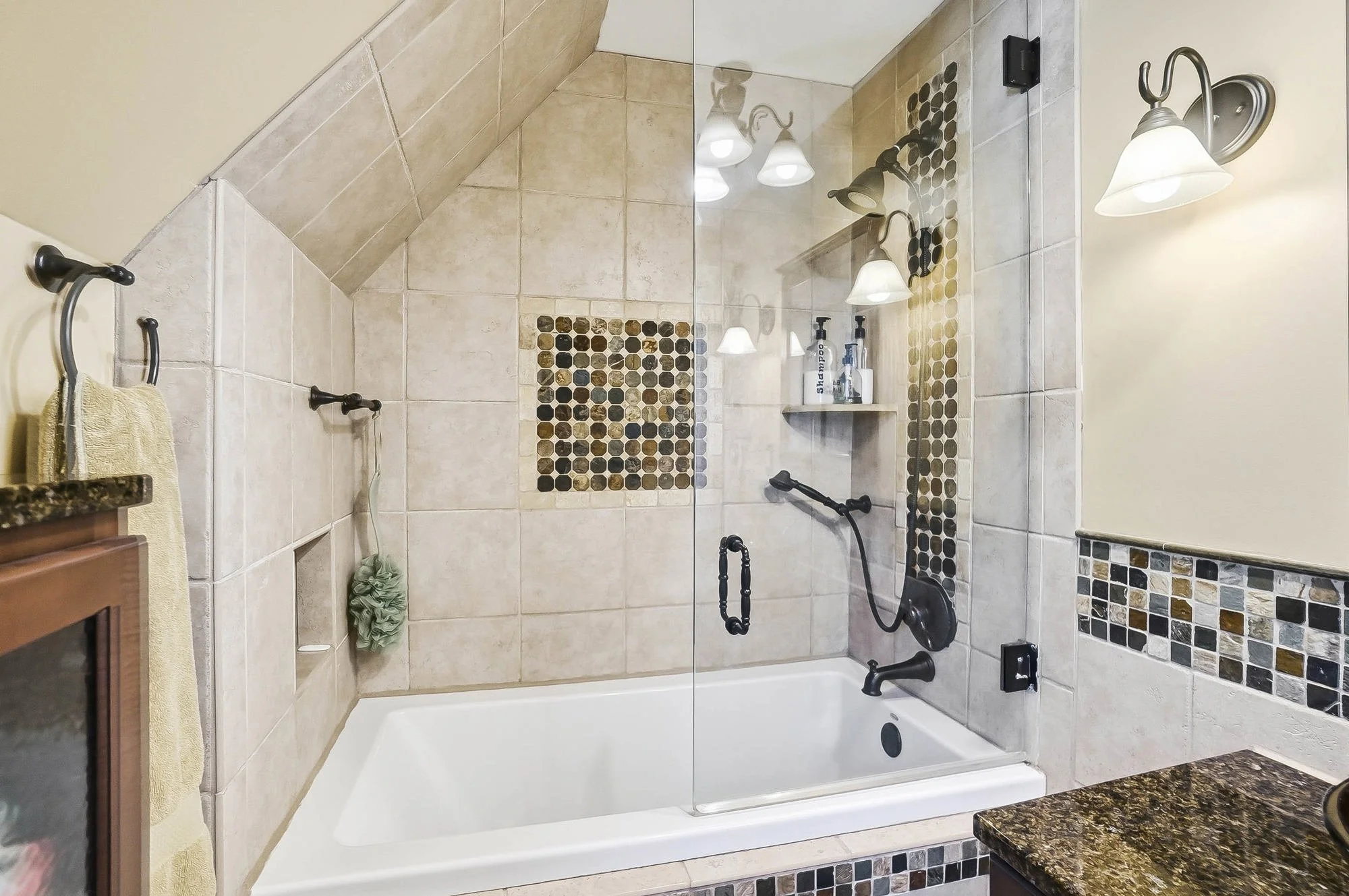 Bathroom with bathtub, tiled walls with decorative mosaic accents, glass shower enclosure, and wall-mounted light fixtures.