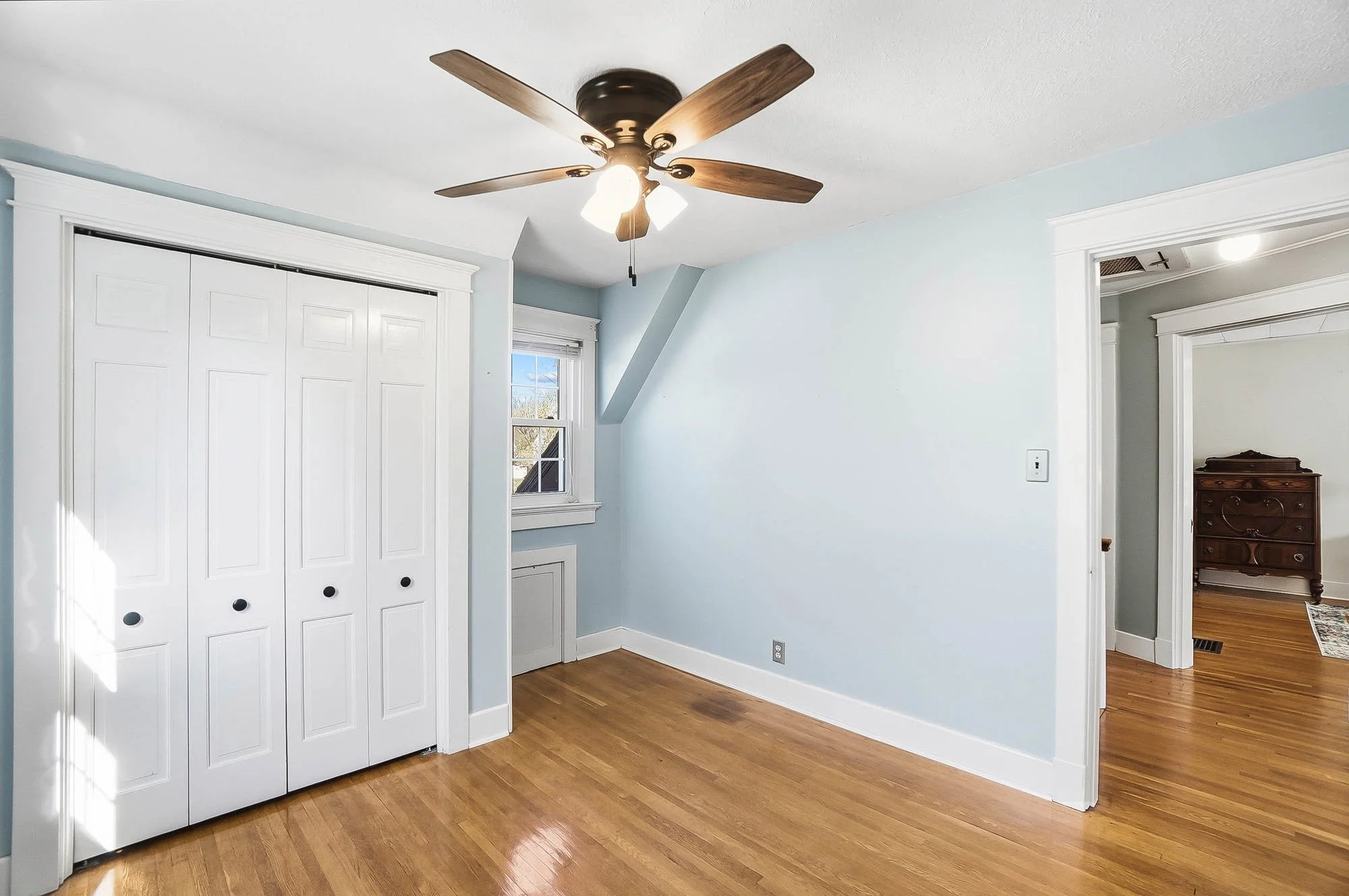 Empty bedroom with hardwood floors, light blue walls, a ceiling fan, a small window, and a white closet door.