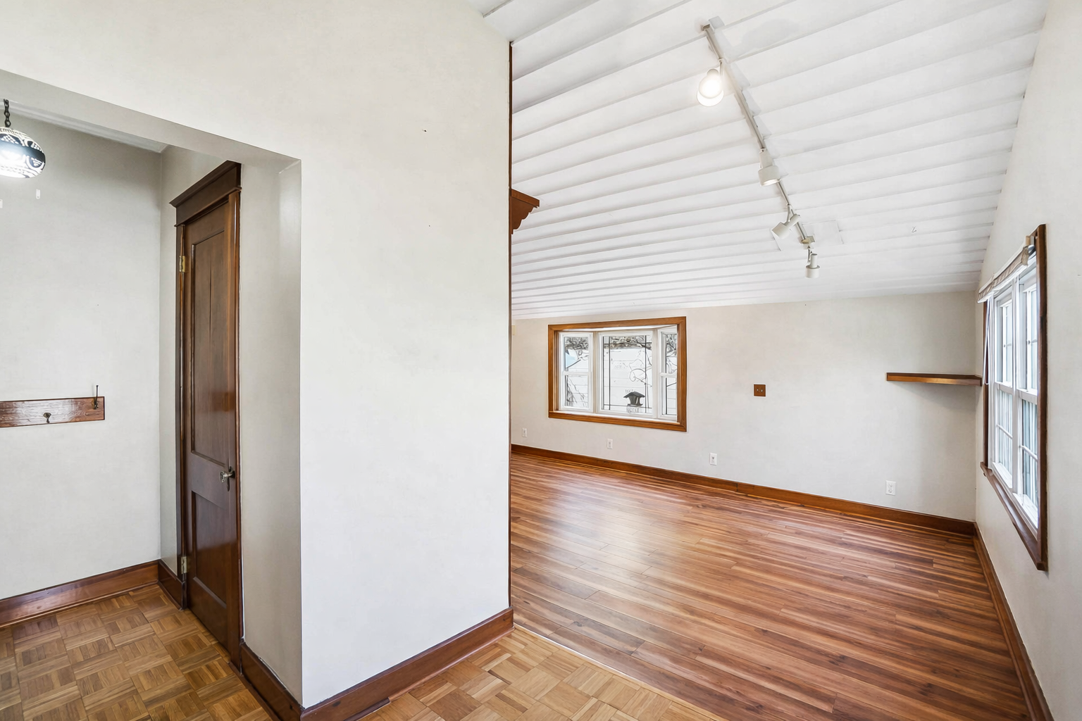 Empty living room with wooden floors, white walls, large window, and ceiling track lighting.