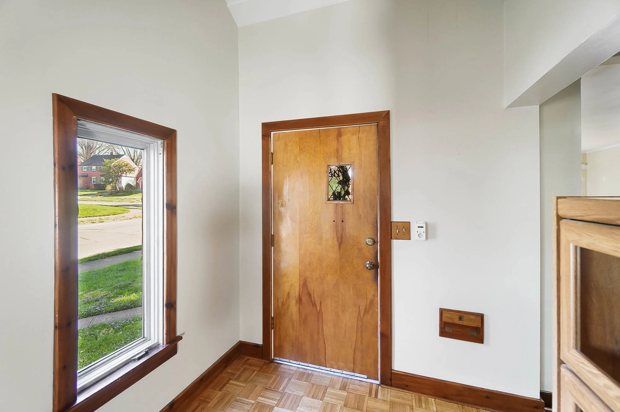 Interior view of a house entryway with a wooden front door that has a small window, a side window showing a lawn and neighboring houses outside, wooden trim around the windows and door, hardwood floor, and a wall-mounted switch and mailbox.