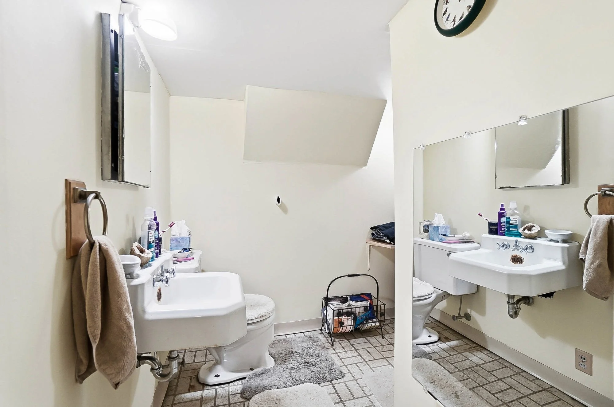 Two white porcelain sinks with towels hanging on wooden racks, a mirror above each sink, and various toiletries on the sinks. A wall clock, a small window, a towel on the floor, and a laundry basket with supplies.