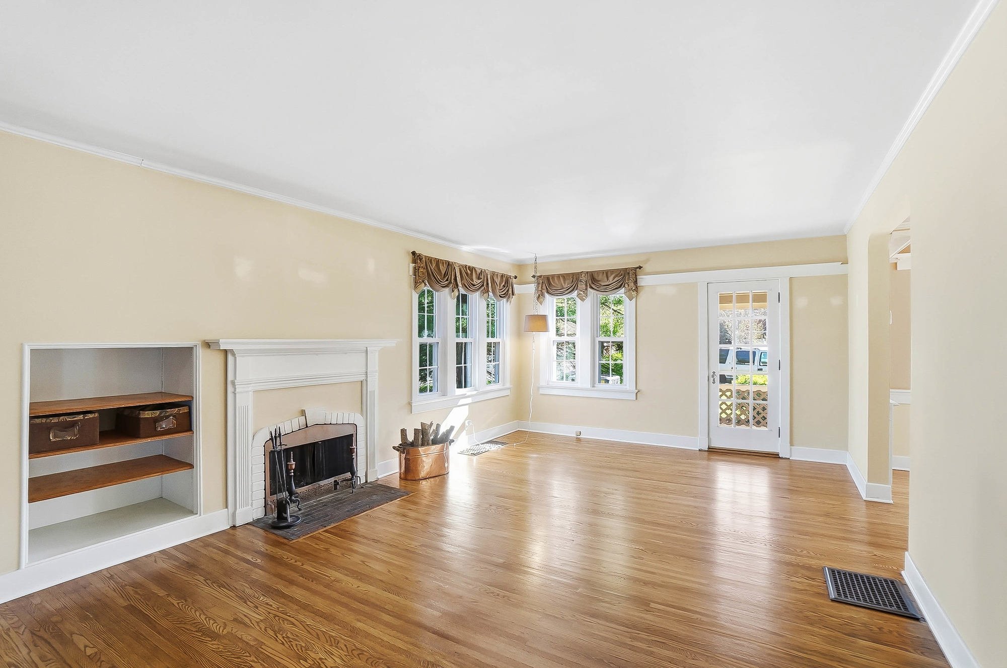 Empty living room with hardwood floors, beige walls, a white fireplace, built-in shelves, large windows with curtains, and a glass door leading outside.