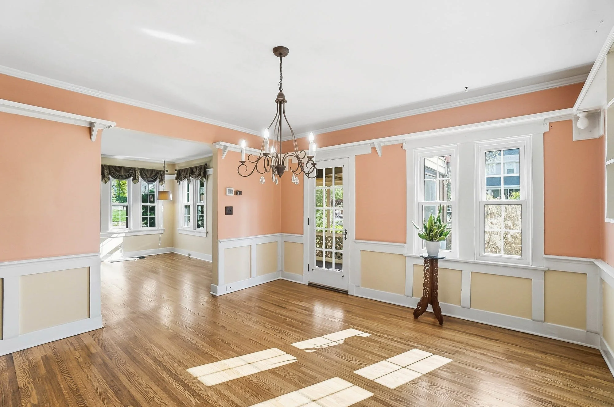 Empty living room with peach and cream walls, hardwood floors, large windows, a glass door, and a chandelier hanging from the ceiling.