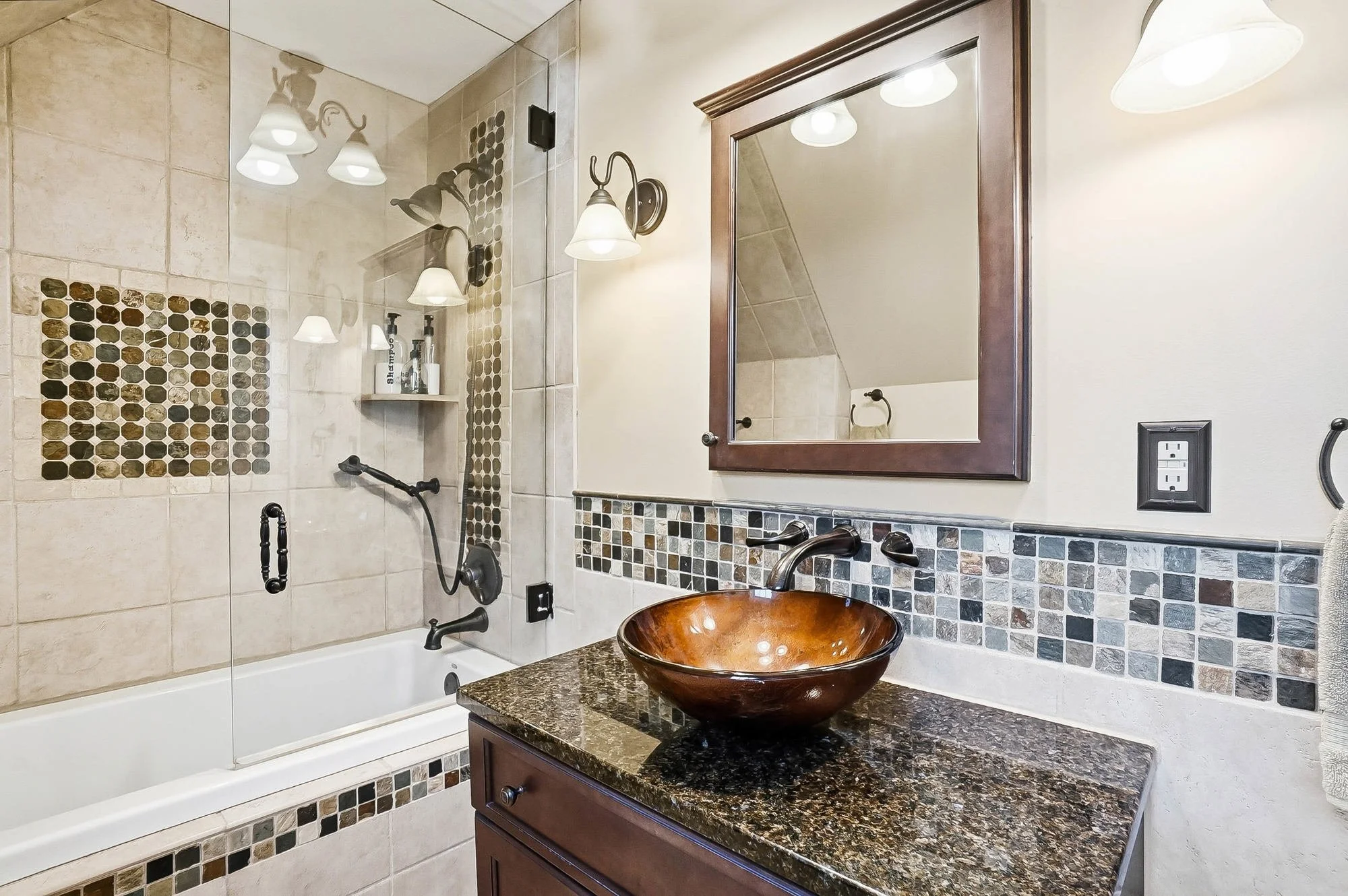 Bathroom with a bathtub and shower, decorated with beige tiles and mosaic accents, a wooden vanity with a granite countertop, and a copper vessel sink with a mirror above.