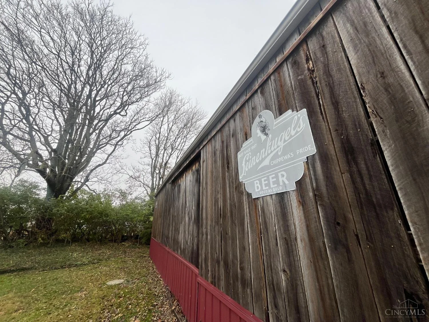 Photo of a wooden building with a gray sign that reads "Leinenkugel's Beer" and shows an image of a person's head, outdoors on a cloudy day with leafless trees in the background.