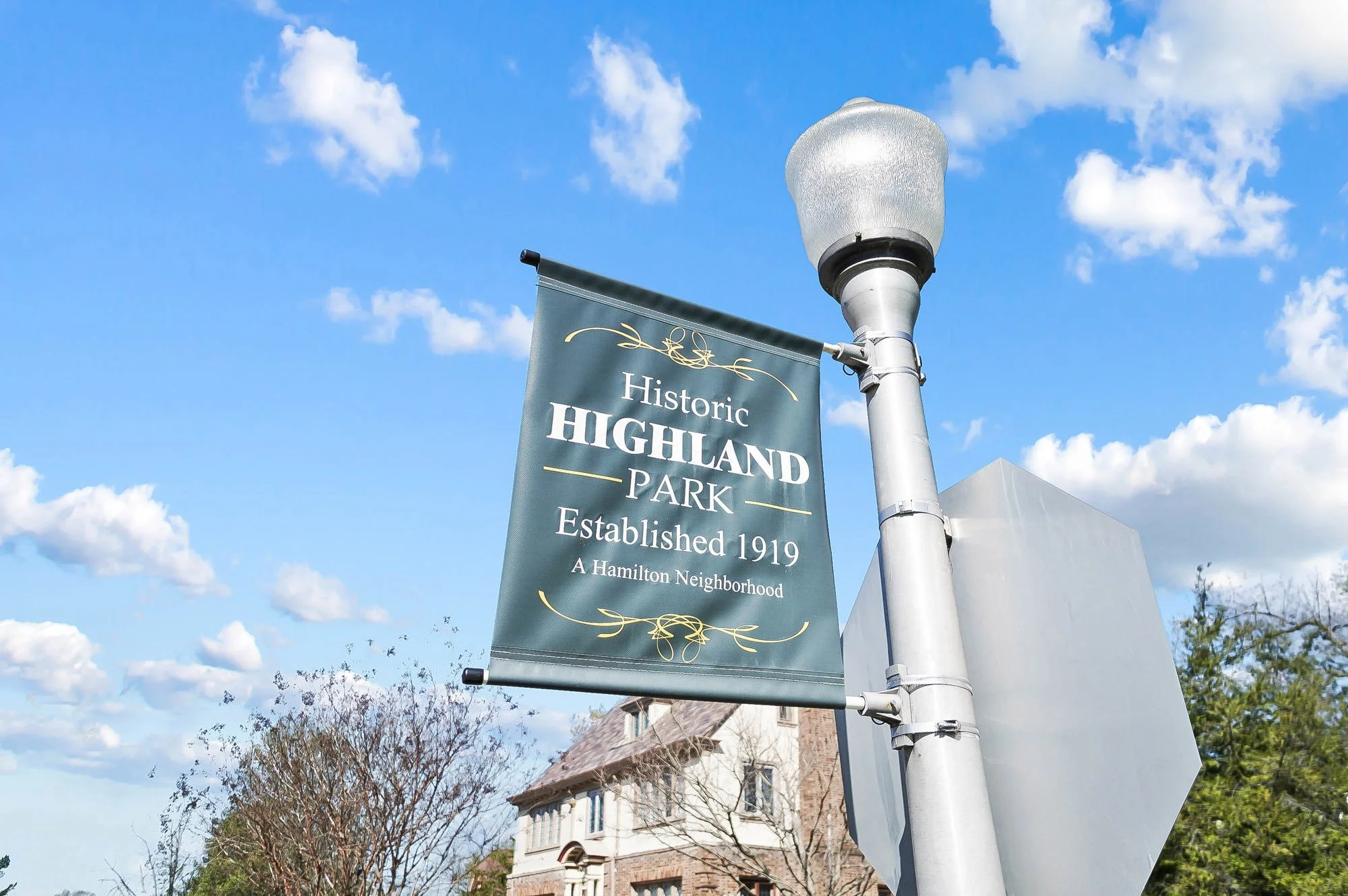 A banner hanging from a street lamp reading "Historic Highland Park Established 1919 A Hamilton Neighborhood" with a house and trees in the background and a blue sky with clouds.