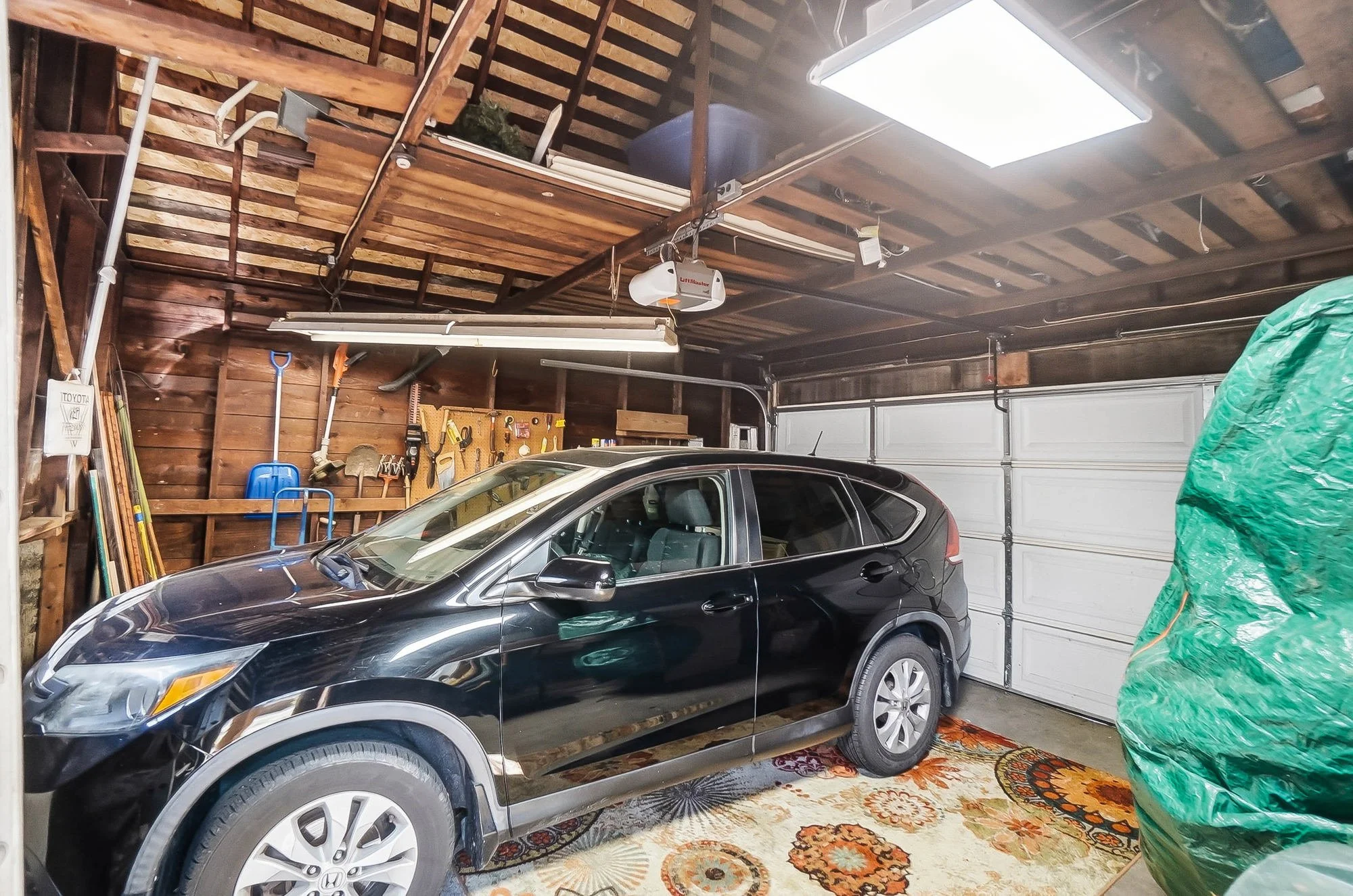 Black SUV parked inside a garage with a wooden ceiling, pegboard with tools on the wall, and a closed white garage door.