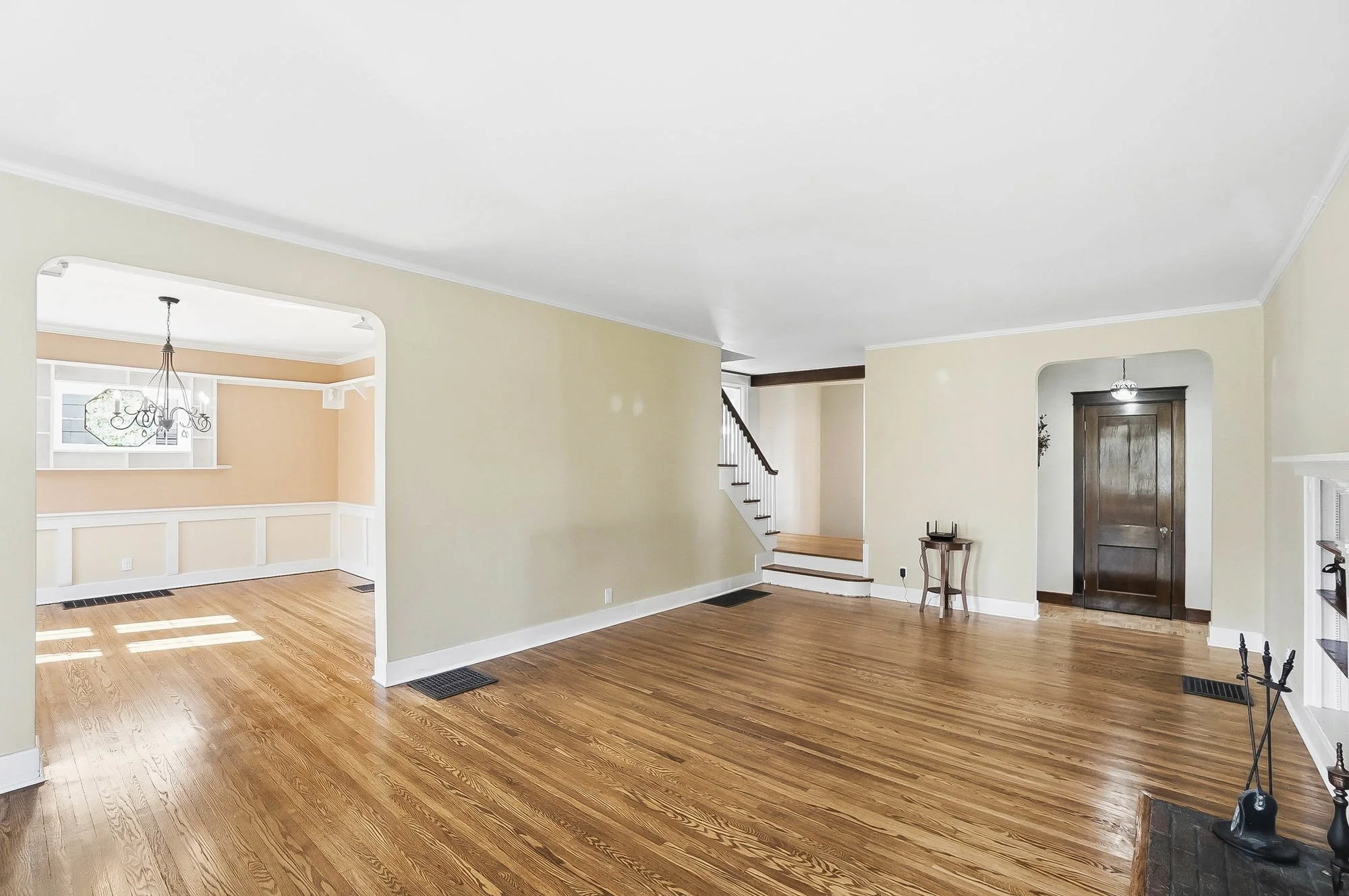Empty living room with hardwood floors, light-colored walls, and dark wooden door, with staircase visible in the background.