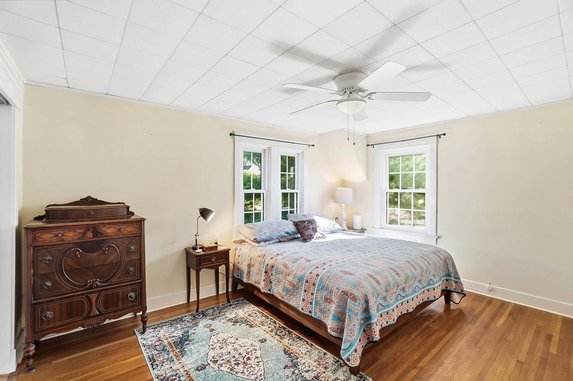 A bedroom with yellow walls, hardwood floors, a ceiling fan, a bed with colorful bedding, two windows with white frames, a nightstand with a lamp and books, an ornate wooden dresser, a small bedside table with a lamp, and a patterned rug.