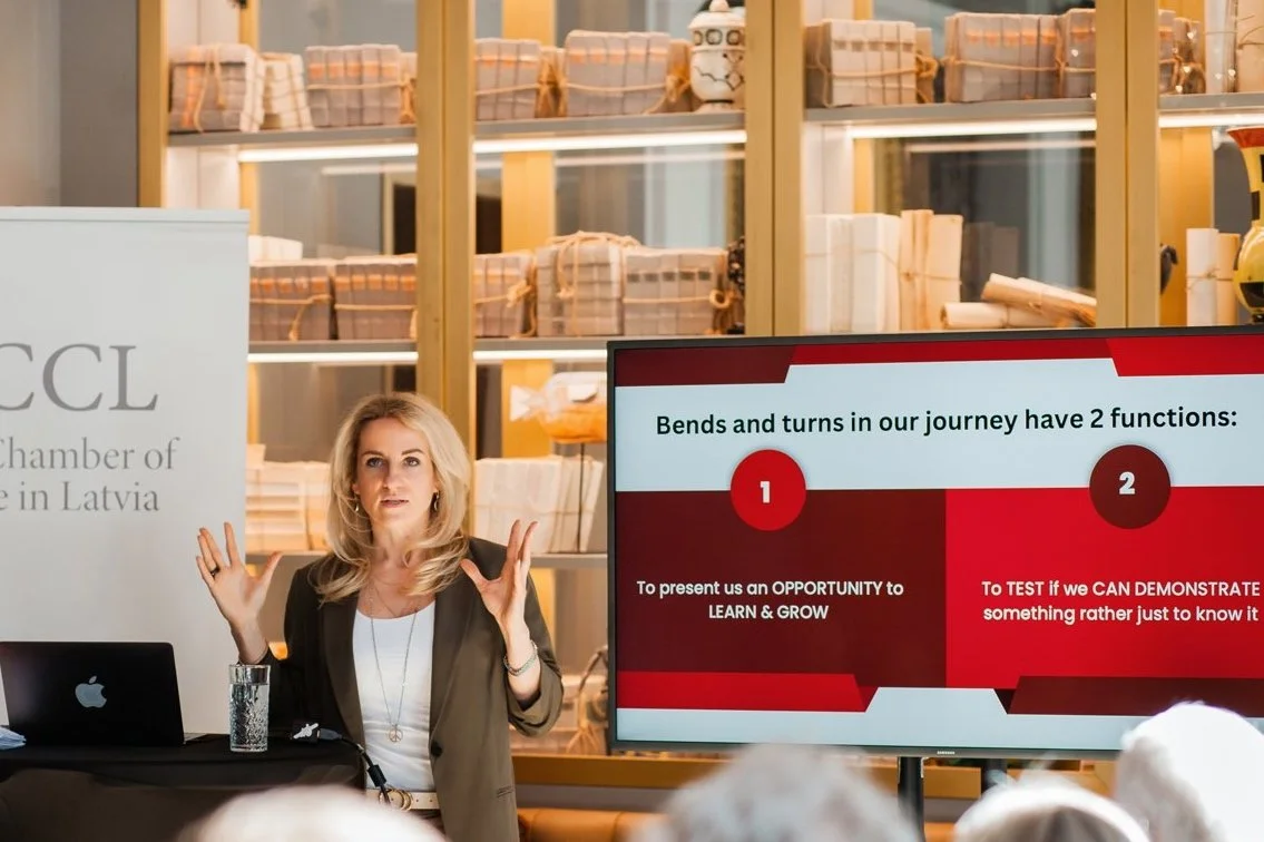 Woman giving a presentation in front of a screen with a slide about the journey's functions, with shelves of books or documents in the background.