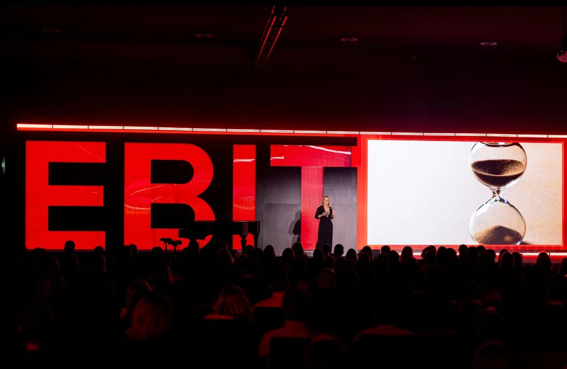 A woman is speaking on stage at a TED event, with the letters 'ERIT' displayed on large red screens and a graphic of an hourglass on the right side.