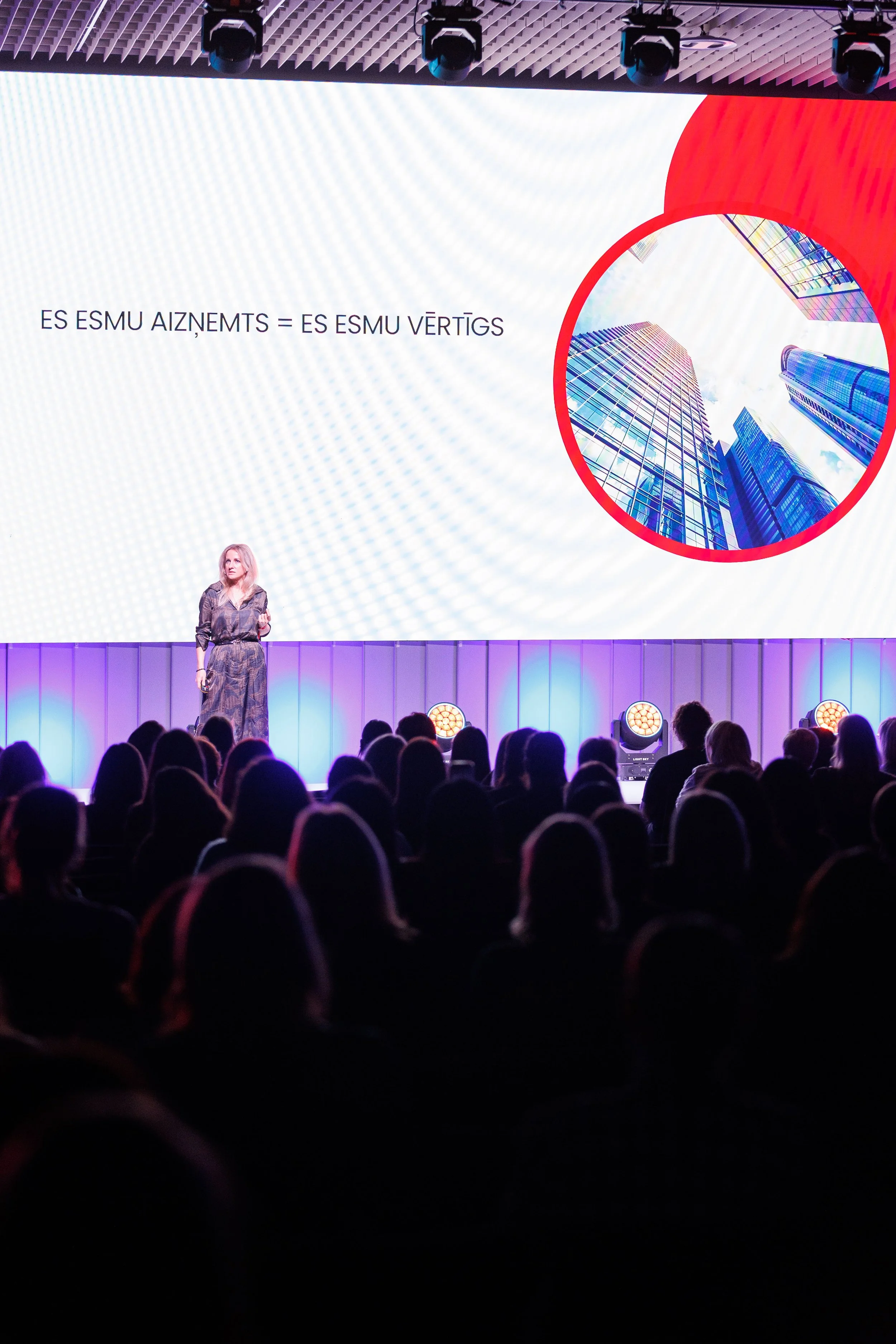 A woman is giving a presentation on stage in front of an audience. The large screen behind her displays an architectural graphic with text in a foreign language and an image of modern skyscrapers.
