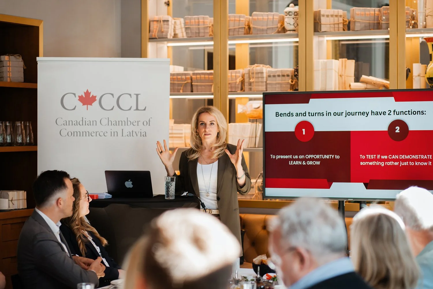 A woman giving a presentation at the Canadian Chamber of Commerce in Latvia event. She is standing next to a display screen showing a slide about the functions of bends and turns in a journey. Several audience members are seated, watching her. There is a banner with the logo and name of the Canadian Chamber of Commerce in Latvia on the left.