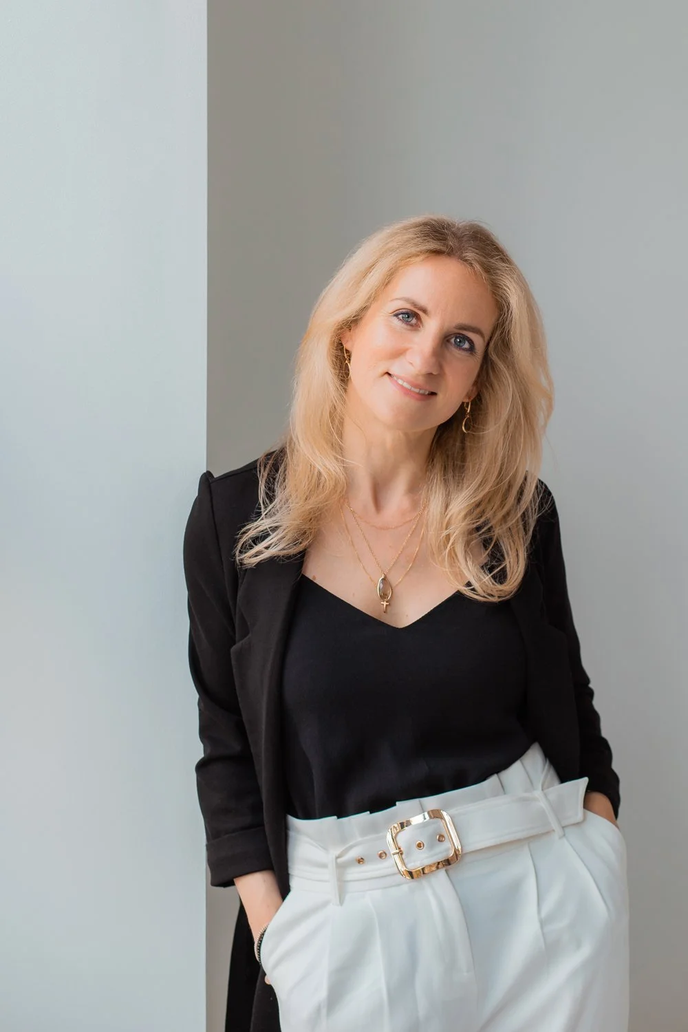 A woman with blonde hair, wearing a black top, white high-waisted pants with a belt, and gold jewelry, standing against a neutral background.