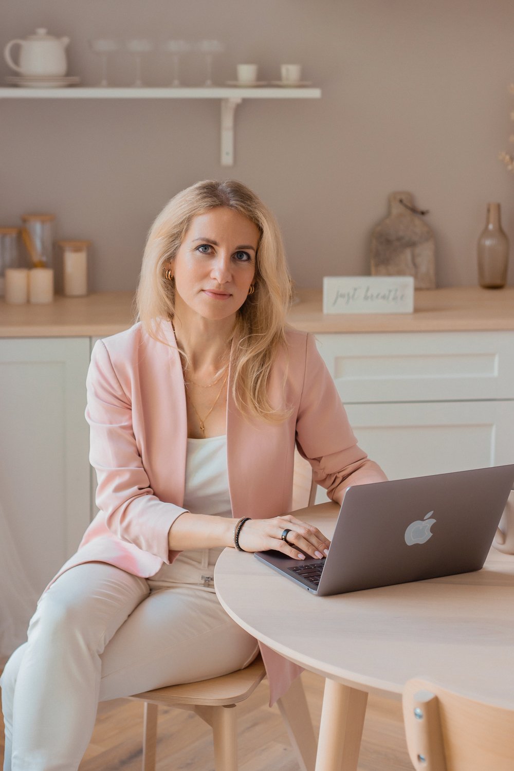 A woman with blonde hair, wearing a pink blazer and white top, sitting at a wooden table with a silver MacBook in a modern, minimalist kitchen or dining area.