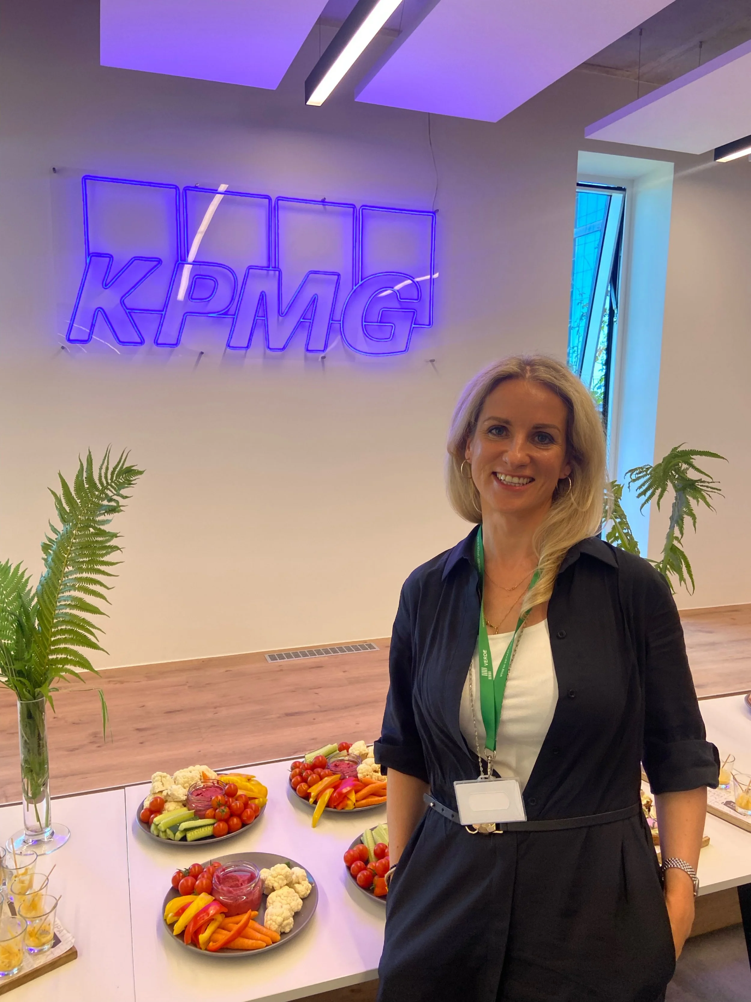 A woman standing in front of a table with fruit platter at a modern office or conference space. There is a neon sign in the background with the text 'KPMG'.