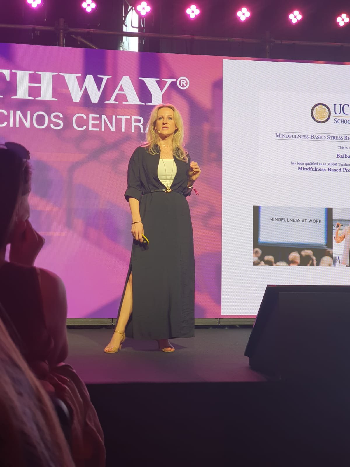 A woman in a black dress and blazer standing on a stage, speaking in front of a large screen with pink and purple colors during a presentation or conference.