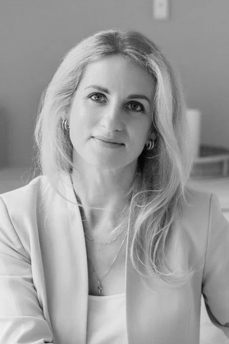 Black and white portrait of a woman with long, wavy hair, wearing earrings, a necklace, and a blazer, sitting indoors.