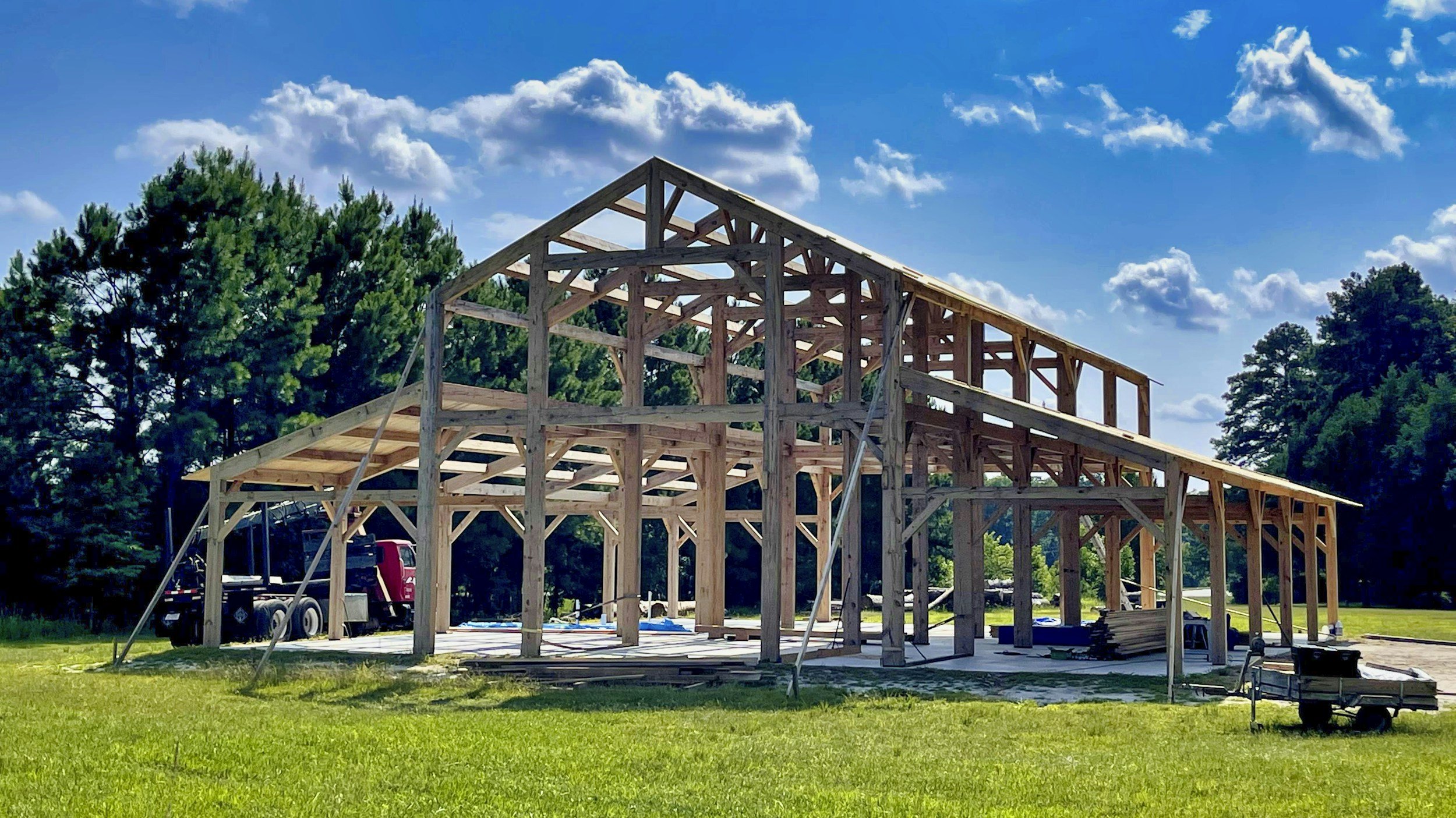 Construction site with a wooden building framework in progress on a grassy area, with trees in the background and blue sky with scattered clouds.