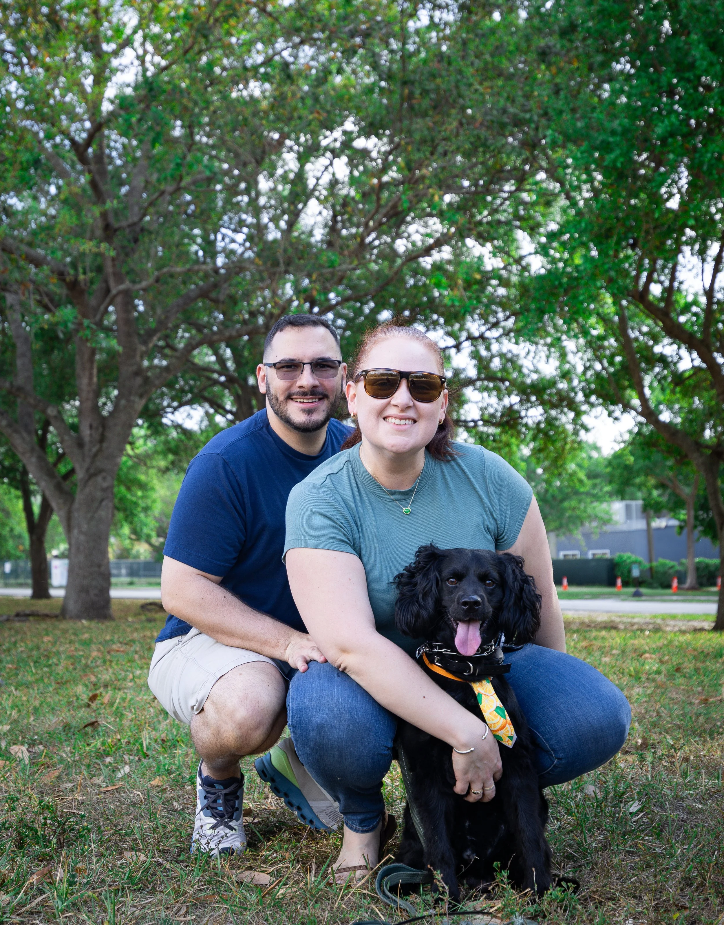 Smiling couple with their black dog in a park with green trees in the background