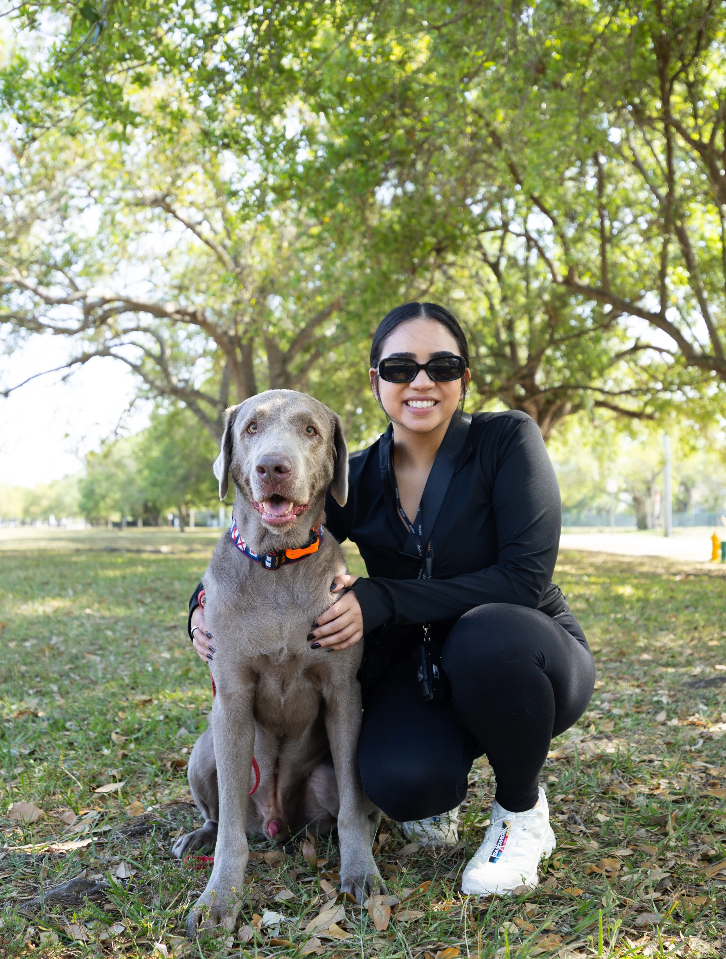 A woman with black hair wearing sunglasses and black athletic clothing, crouching outdoors with a smiling dog in a park with green trees and grass.