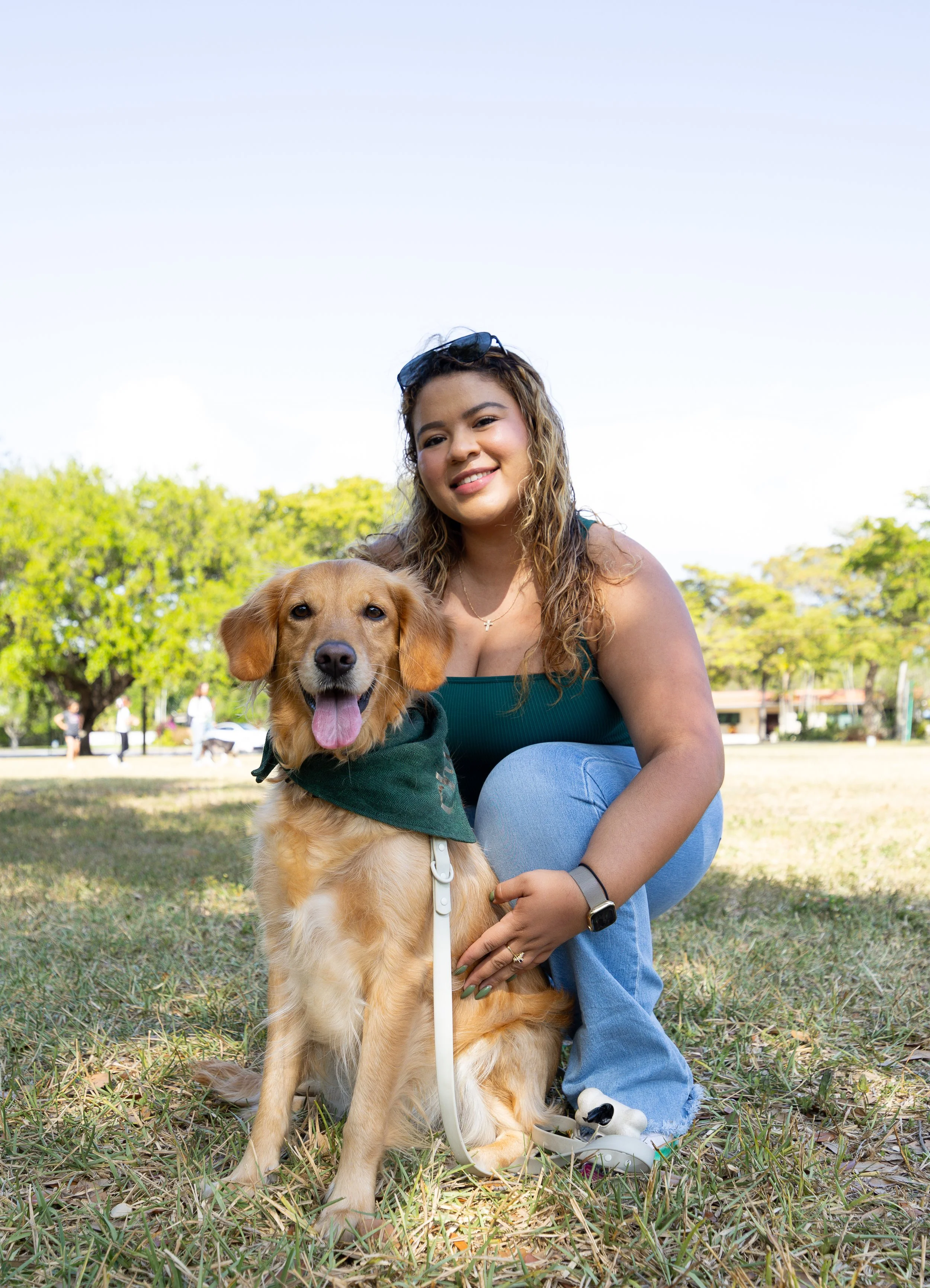 A woman kneeling on grass in a park with her golden retriever dog, both smiling. The woman has curly hair and is wearing a green tank top, blue jeans, sunglasses on her head, and a watch on her wrist. The dog has a green bandana around its neck and looks happy with its tongue out. There are trees and other people in the background.
