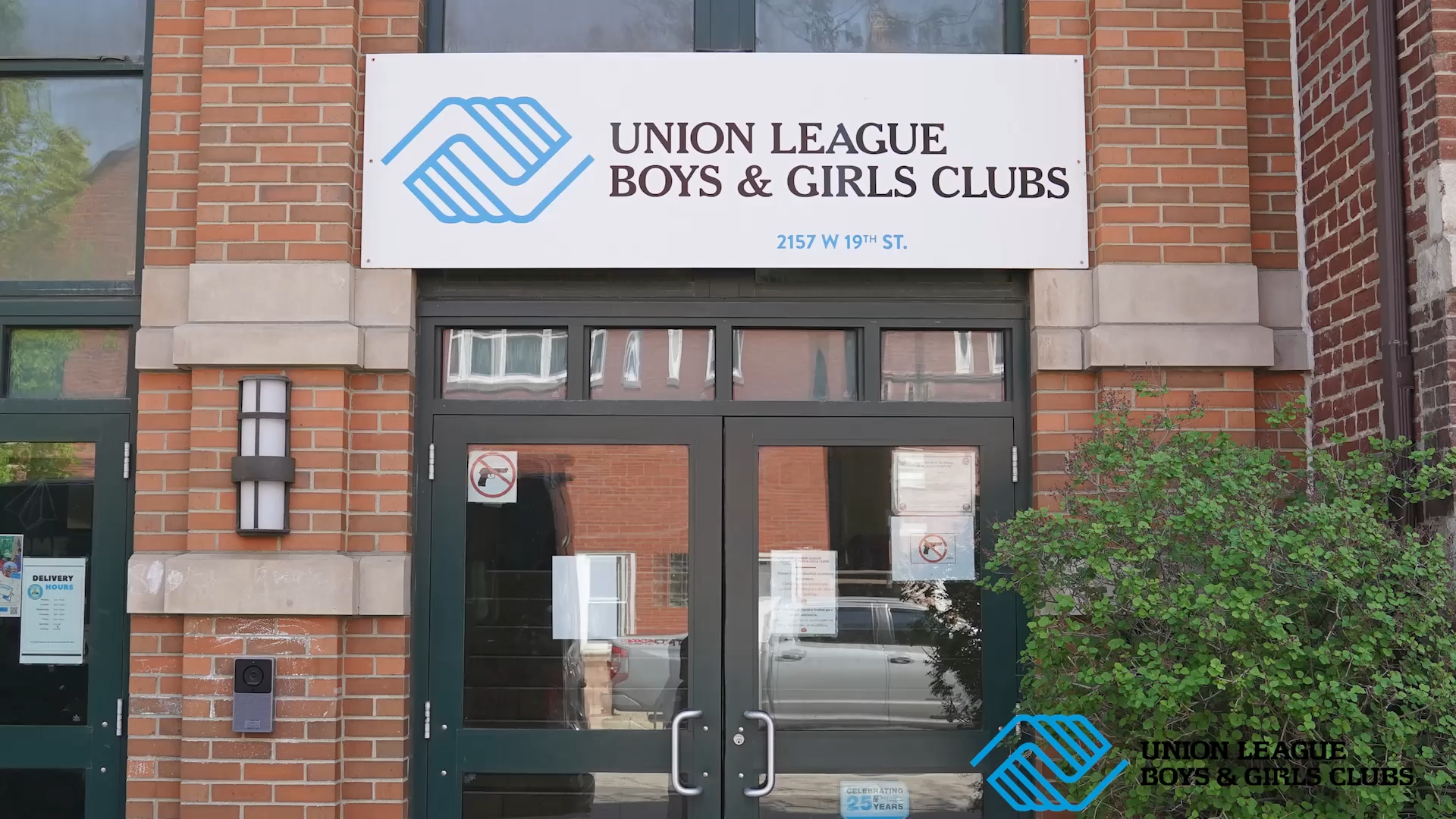 Building entrance with a Union League Boys & Girls Clubs sign above the door. The sign features a blue logo with two shaking hands and the club's name in black text. The address 2157 W 19th St. is printed on the sign. The entrance has glass double do