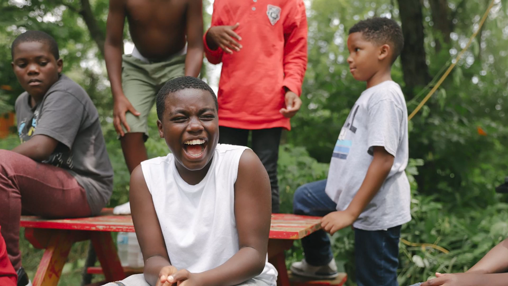 Group of children outdoors, one girl laughing and two boys standing nearby, green trees in the background.