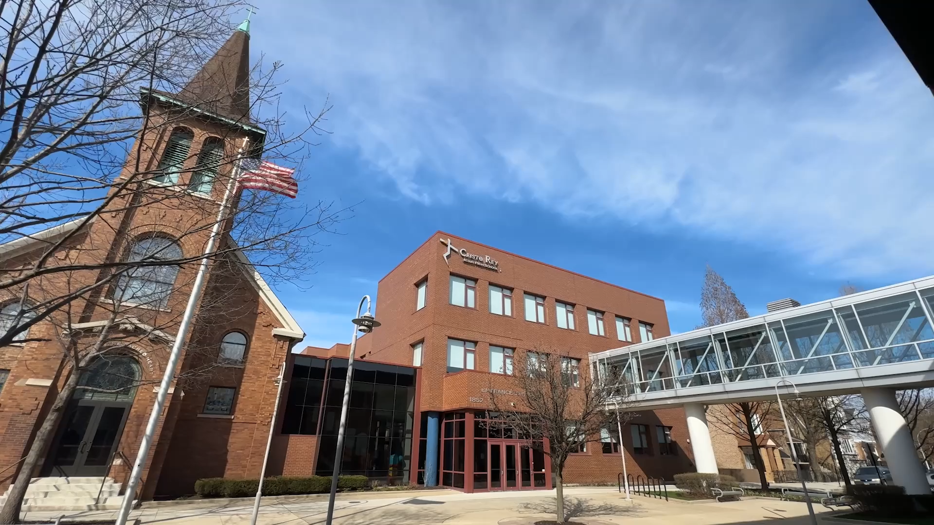 A brick church with a tall steeple and an American flag, beside a modern brick building with a sign that reads 'Cristo Rey Community Center'. A glass pedestrian bridge connects the two structures, with trees and street lamps in the scene under a part