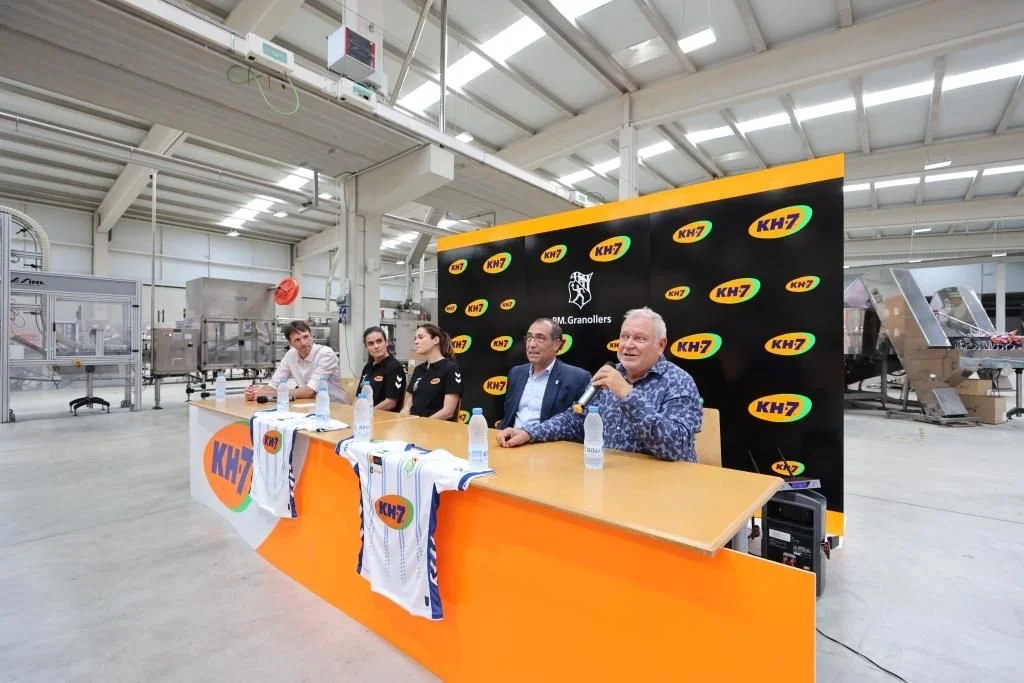 Cuatro personas sentadas en una mesa de presentación en una fábrica o planta de producción, con un fondo negro con logos de KH7 y GM Granollers, y una de ellas hablando con micrófono.