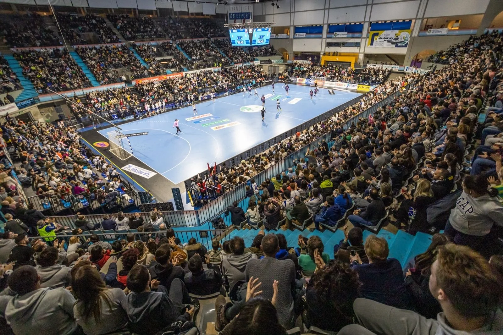 Vista del interior de un estadio durante un partido de balonmano, con espectadores en las gradas y jugadores en la cancha.