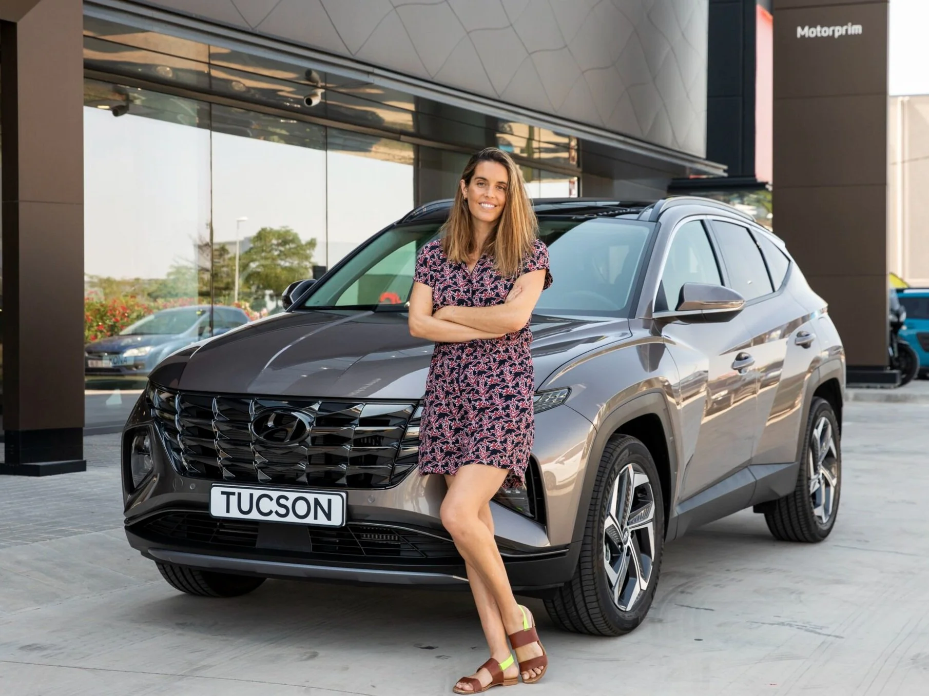 Mujer sonriendo y de brazos cruzados junto a un auto Hyundai Tucson gris en un concesionario de autos.