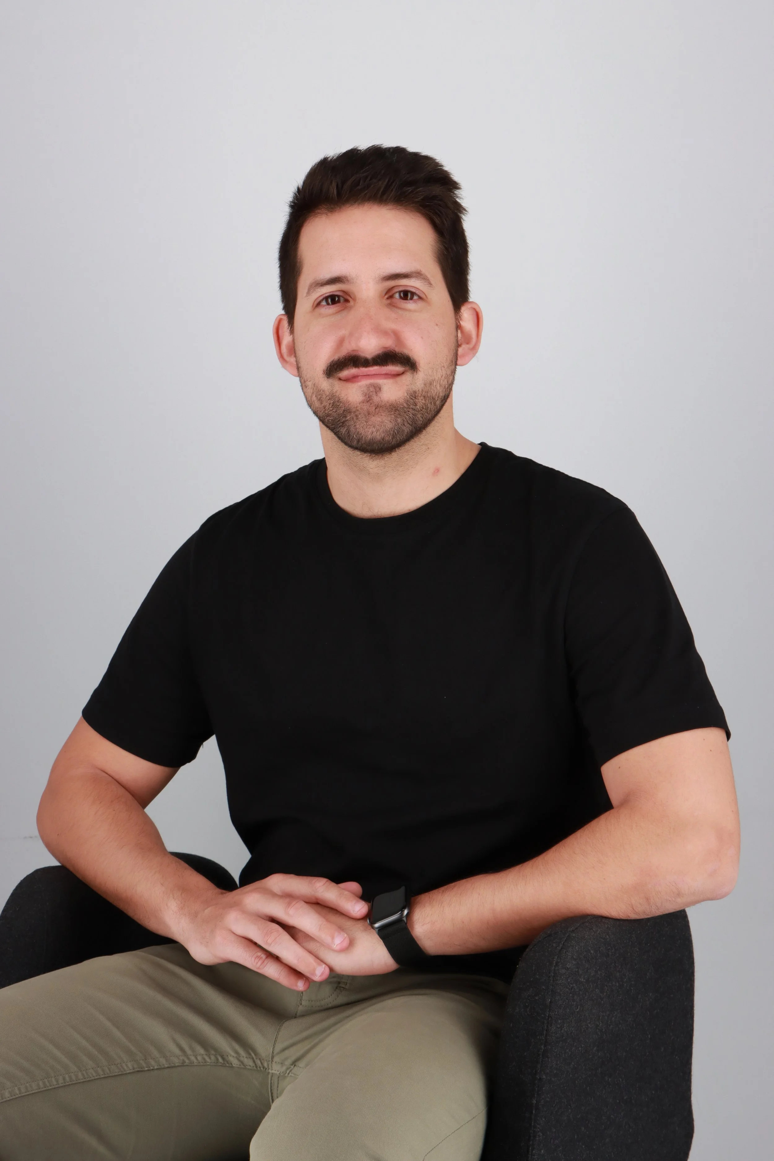 Retrato de un hombre con barba y bigote, con cabello oscuro, usando camiseta negra y pantalones color caqui, sentado en una silla gris, con fondo gris claro.