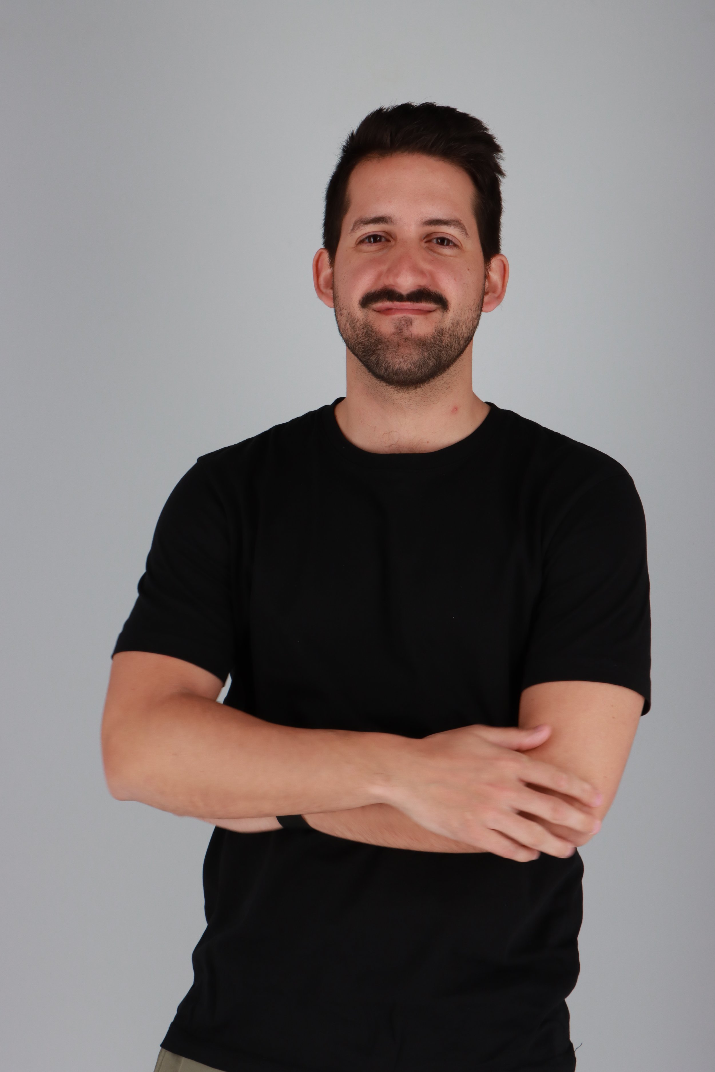 Hombre con barba, cabello oscuro y camiseta negra, mirando a la cámara, de fondo gris