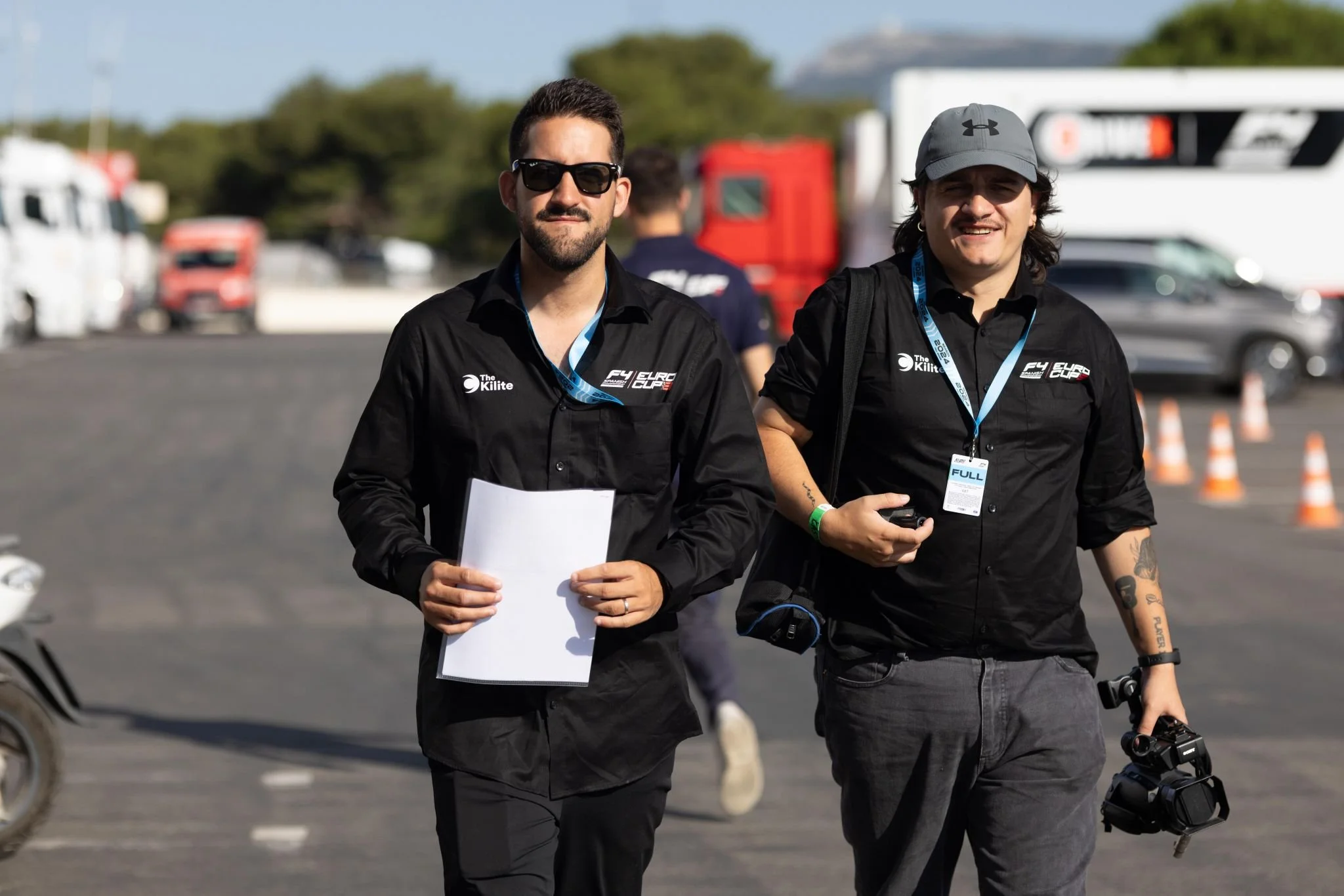 Dos hombres caminando en un área de carreras, llevando equipos de fotografía y papeles, con vehículos y conos de tráfico en el fondo.