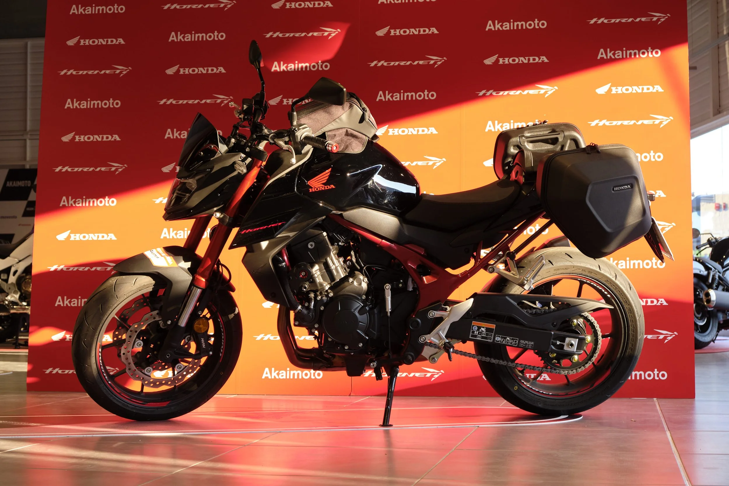Motocicleta negra de Honda en exhibición con fondo rojo y logotipos de marcas como Honda, Akaiomoto y Hornet.