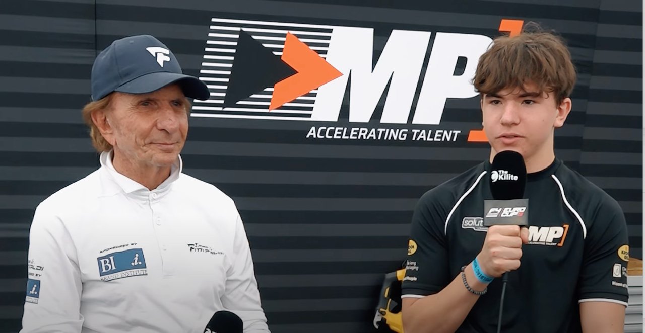 Un joven piloto de carreras con micrófono en un evento de racing, de fondo un cartel que dice 'IMP Accelerating Talent' y un hombre mayor con gorra observándolo