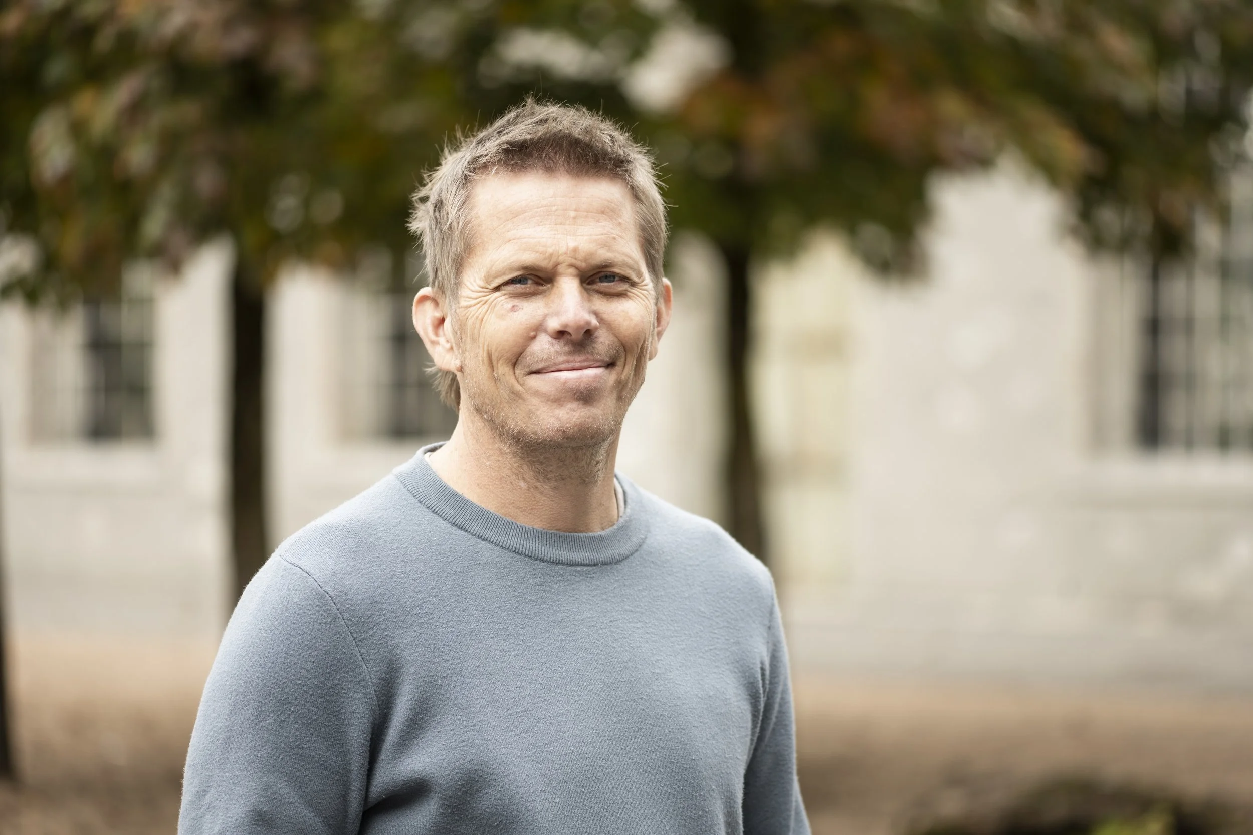 Ein Mann mit hellbraunen Haaren trägt einen grauen Pullover im Freien vor einem Baum und einem Gebäude.