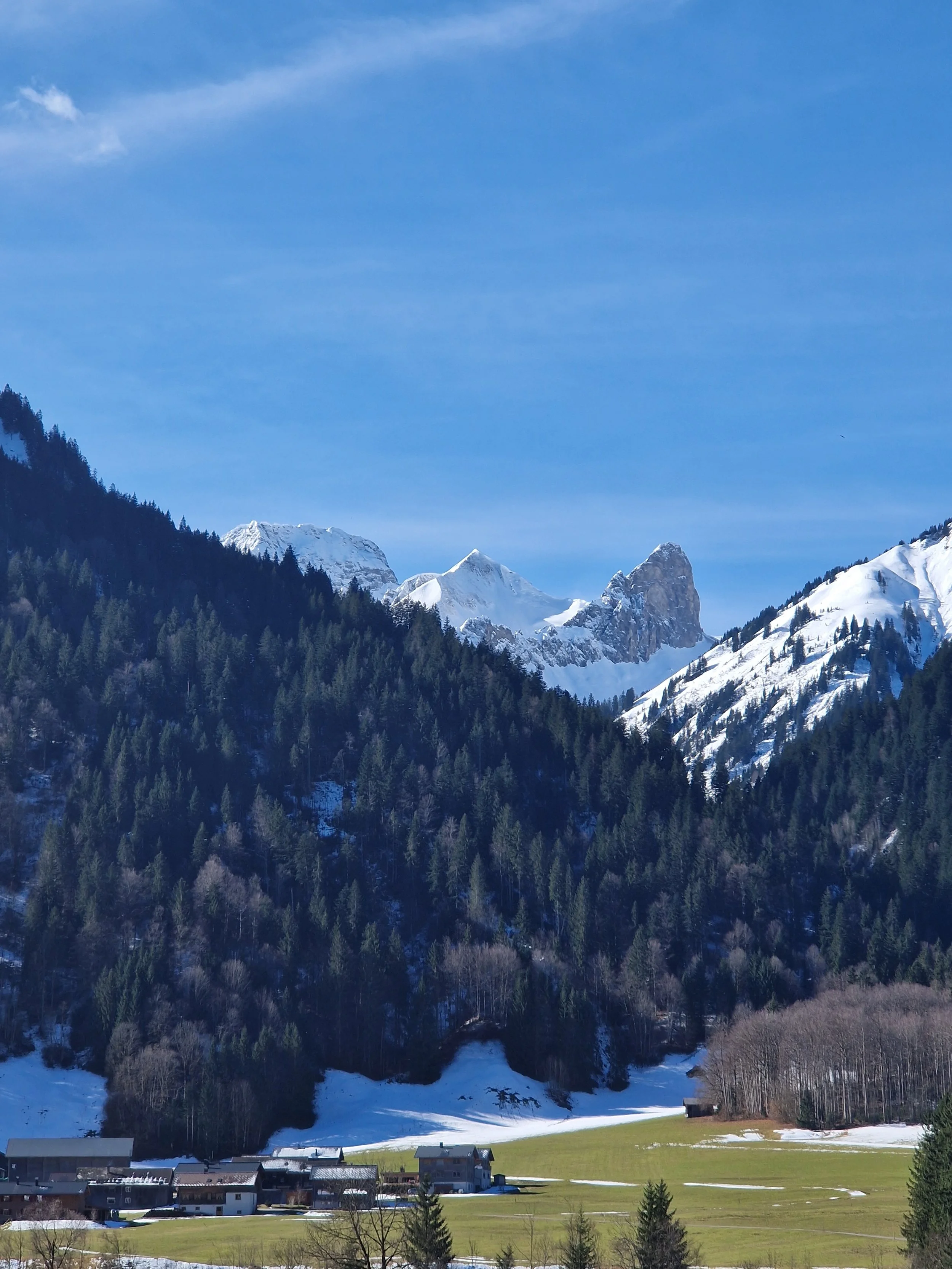 Berge mit Schneefeldern, Wälder und eine kleine Ortschaft in einer Talebene, sonniger Tag