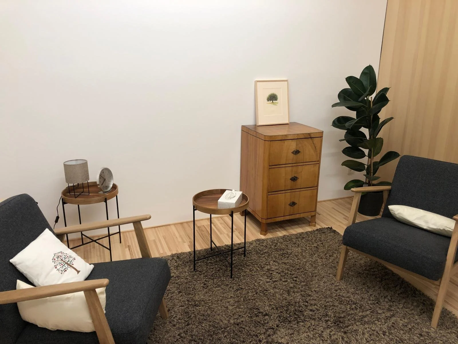Sicht auf ein Wohnzimmer mit zwei dunklen Sofas mit weißen Kissen, zwei runden Holztischen, einer kleinen Holzkommode mit einer kleinen Wandbildrahmen und einer großen Topfpflanze in einer Ecke, auf einem braunen Teppich auf Holzboden.